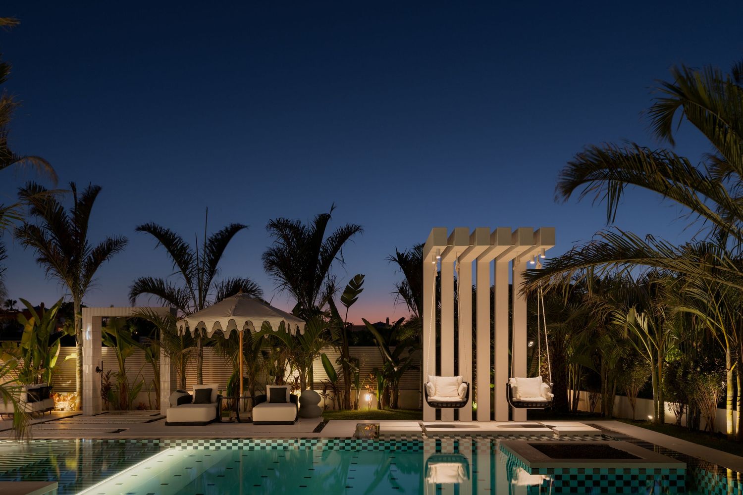 Dramatic nighttime view of white architectural columns and pergola beside a glowing pool surrounded by tropical palm trees.