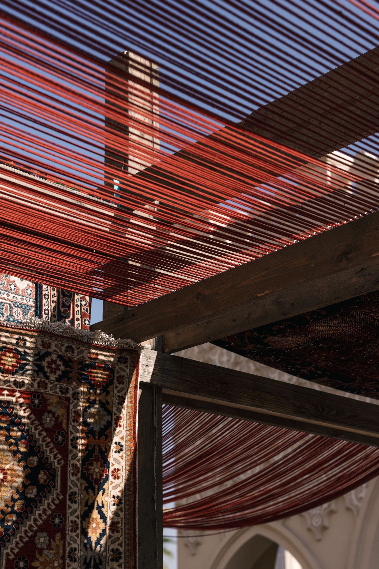 Red threads stretched across a wooden pergola ceiling above hanging traditional Persian carpets in sunlight.