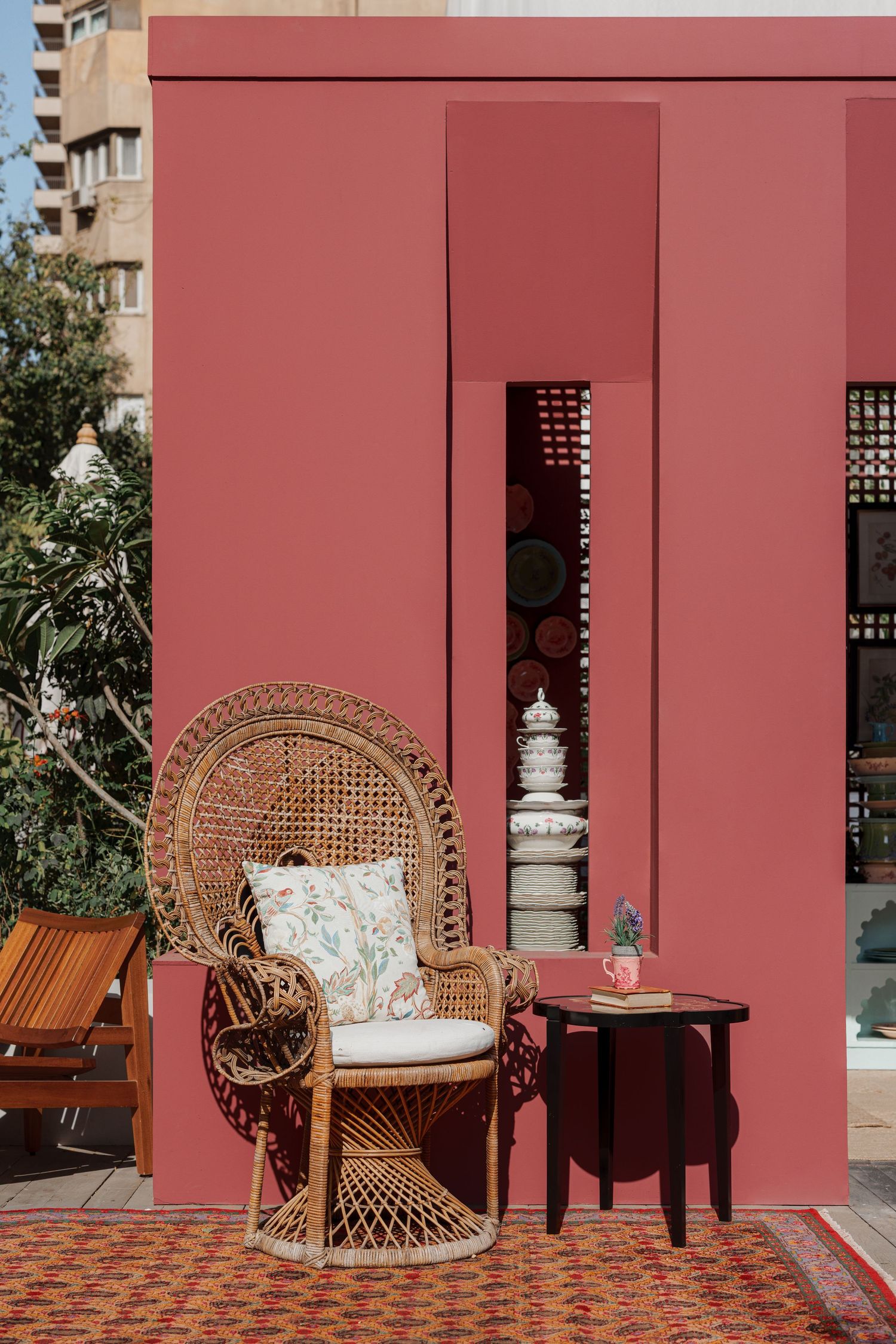 Wicker peacock chair and small table against a terracotta pink building facade outdoors.