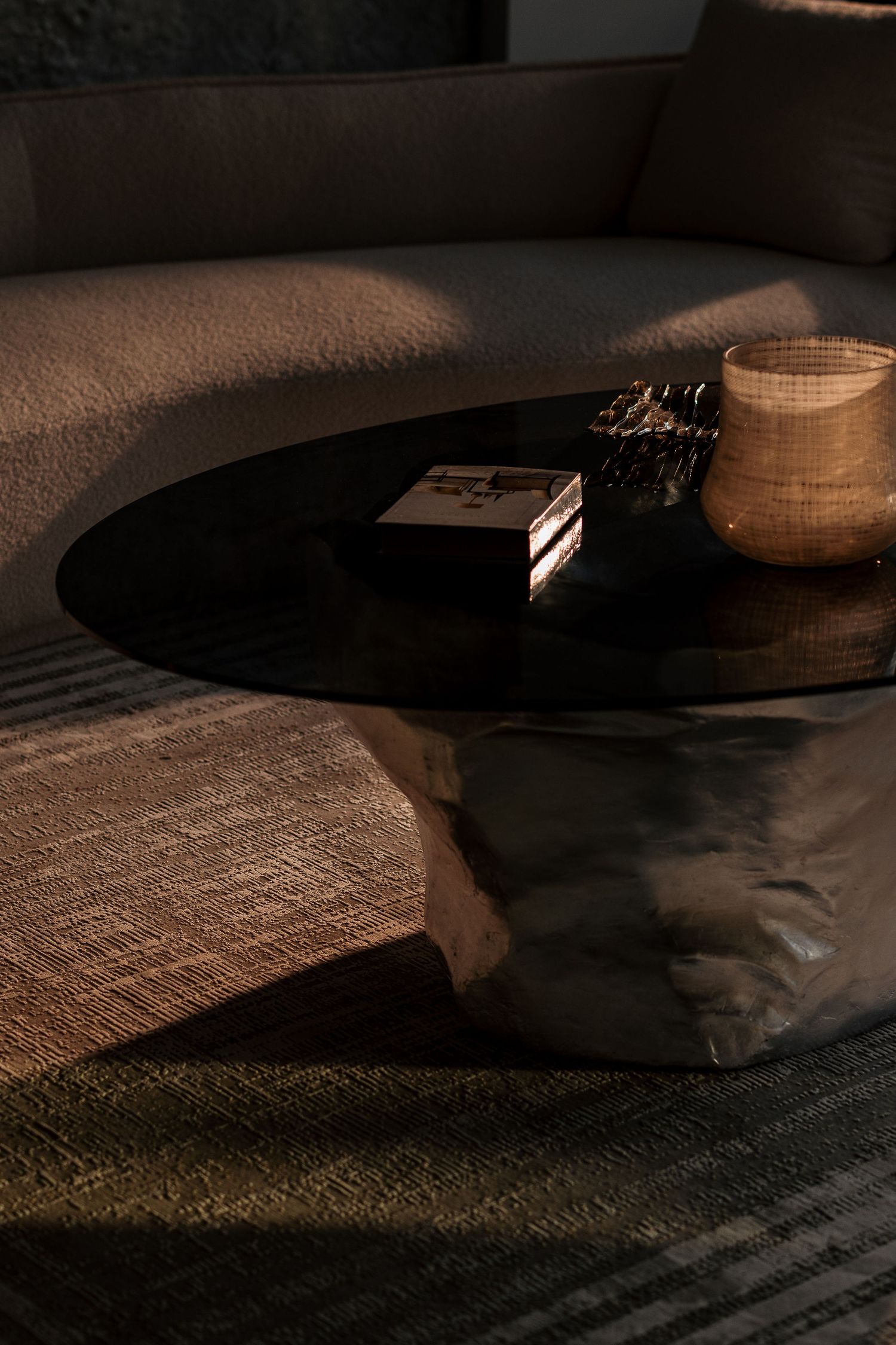 Dark round glass coffee table with decorative objects atop textured stone base beside curved beige sofa on woven rug.
