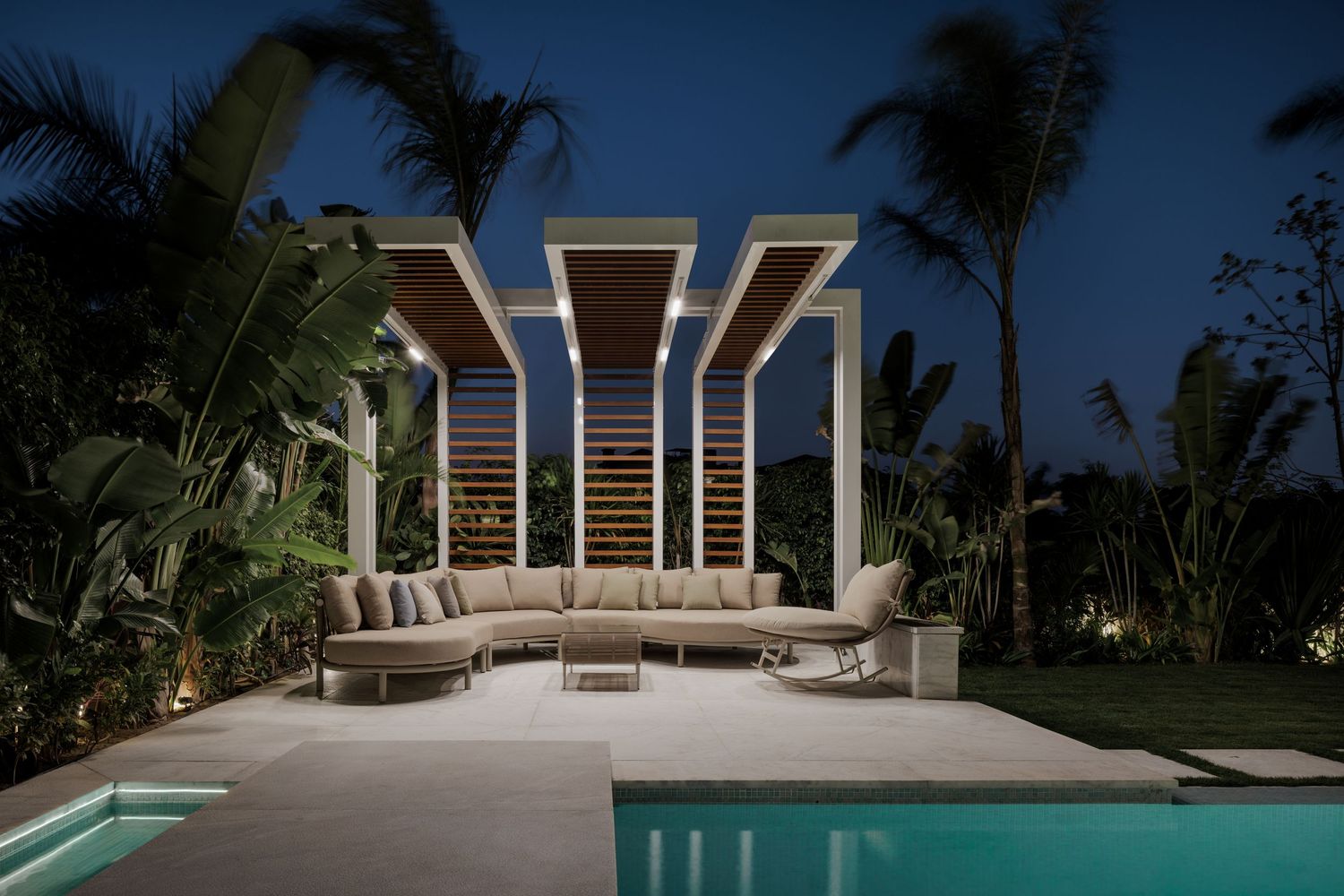 Sophisticated outdoor lounge area at night with sectional sofa under a slatted pergola beside a glowing pool.