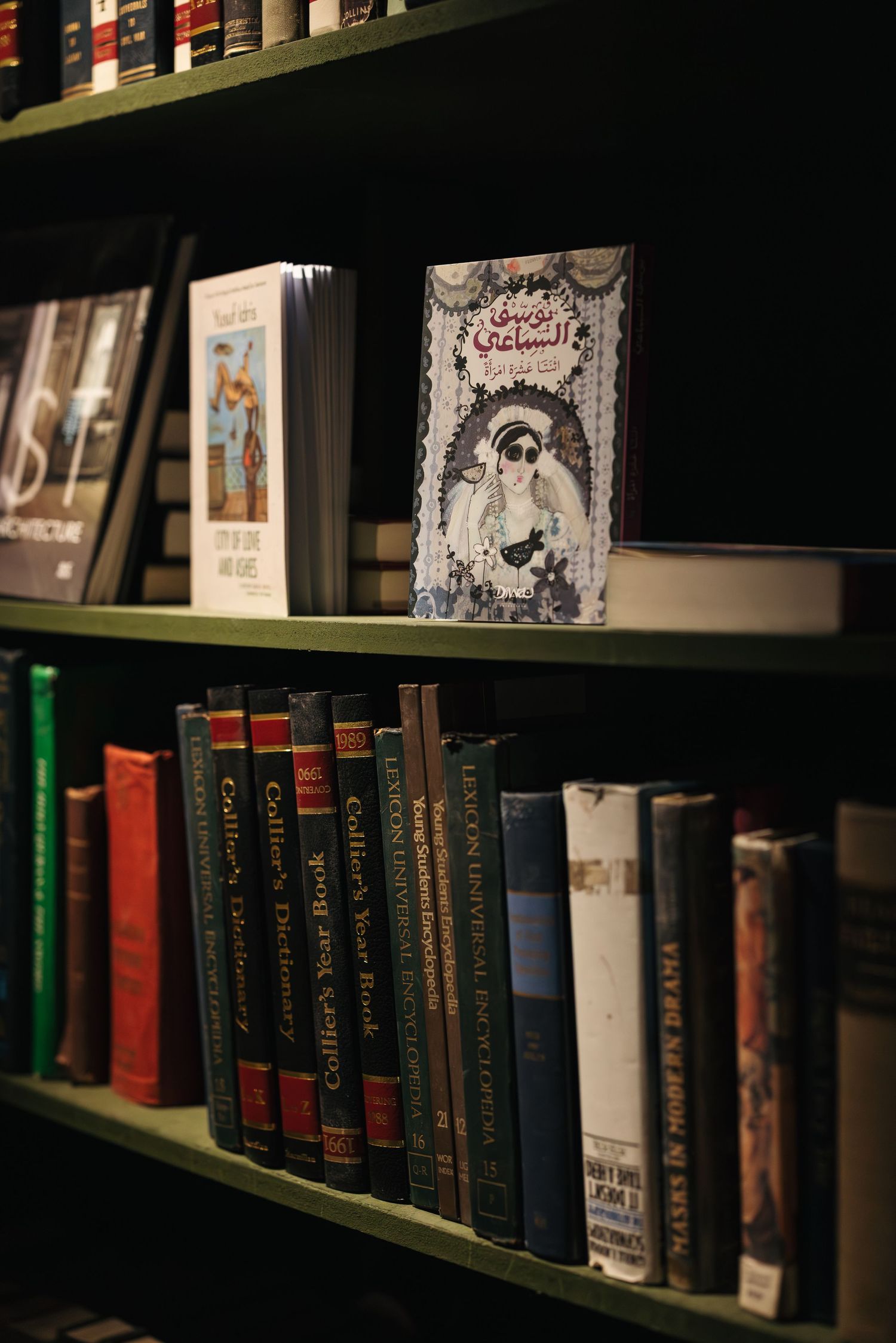 Green bookshelf displaying Arabic illustrated book cover among Collier encyclopedia and architecture volumes.