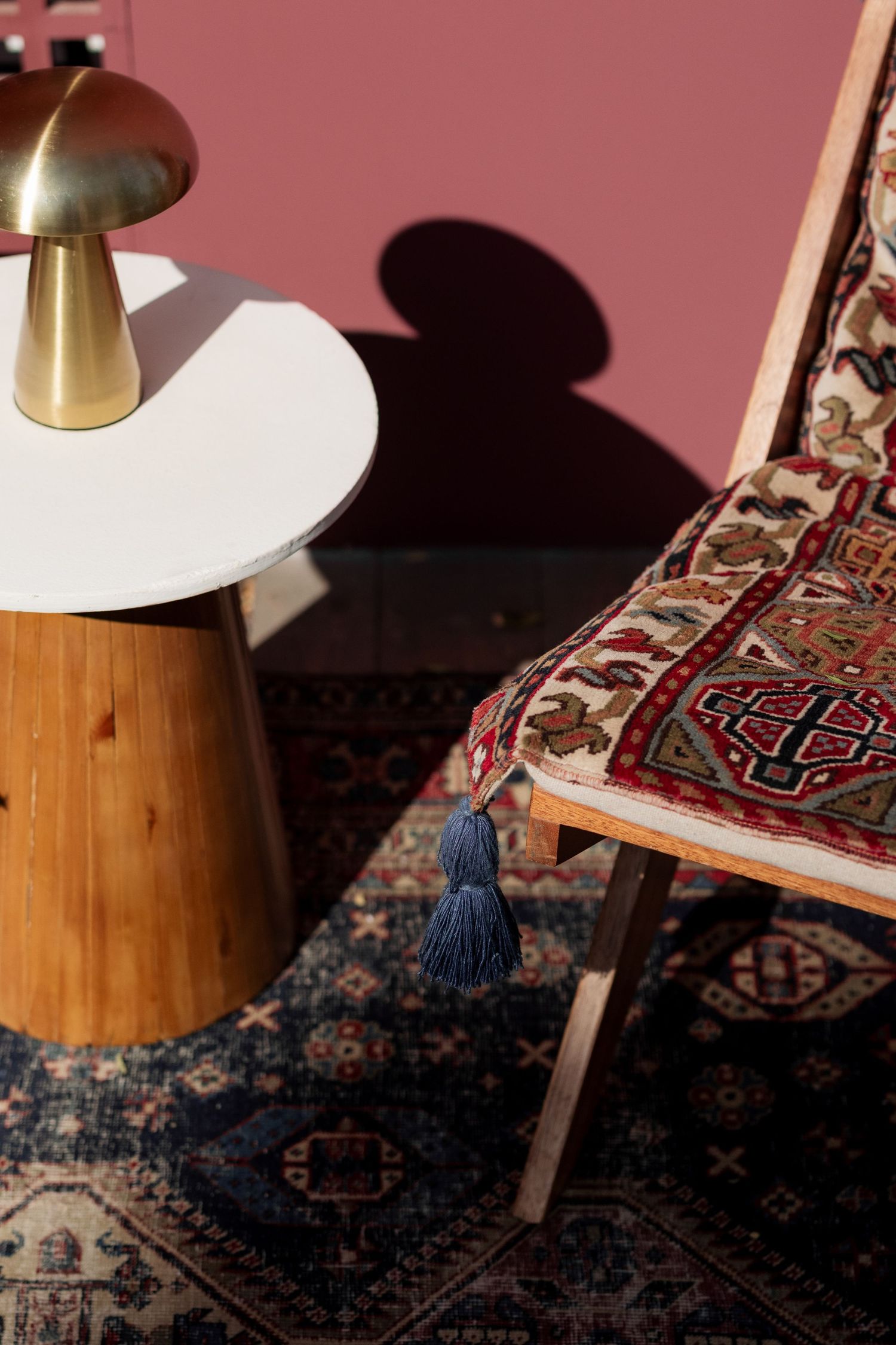 Wooden chair with kilim textile cushion and blue tassel beside a mushroom brass lamp on a round table.