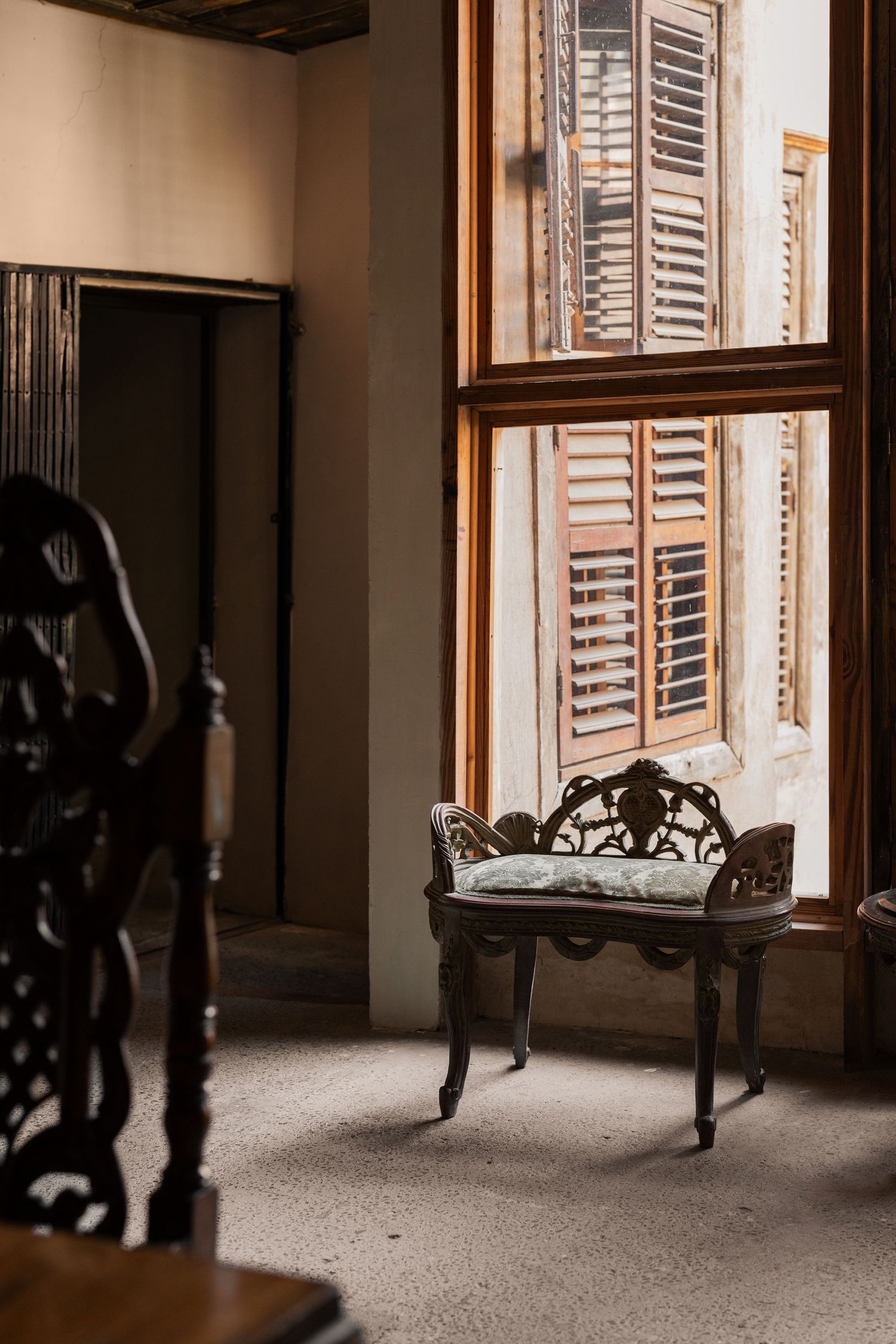 Vintage ornate iron chair beside large wooden shuttered window in a sunlit classical interior room.
