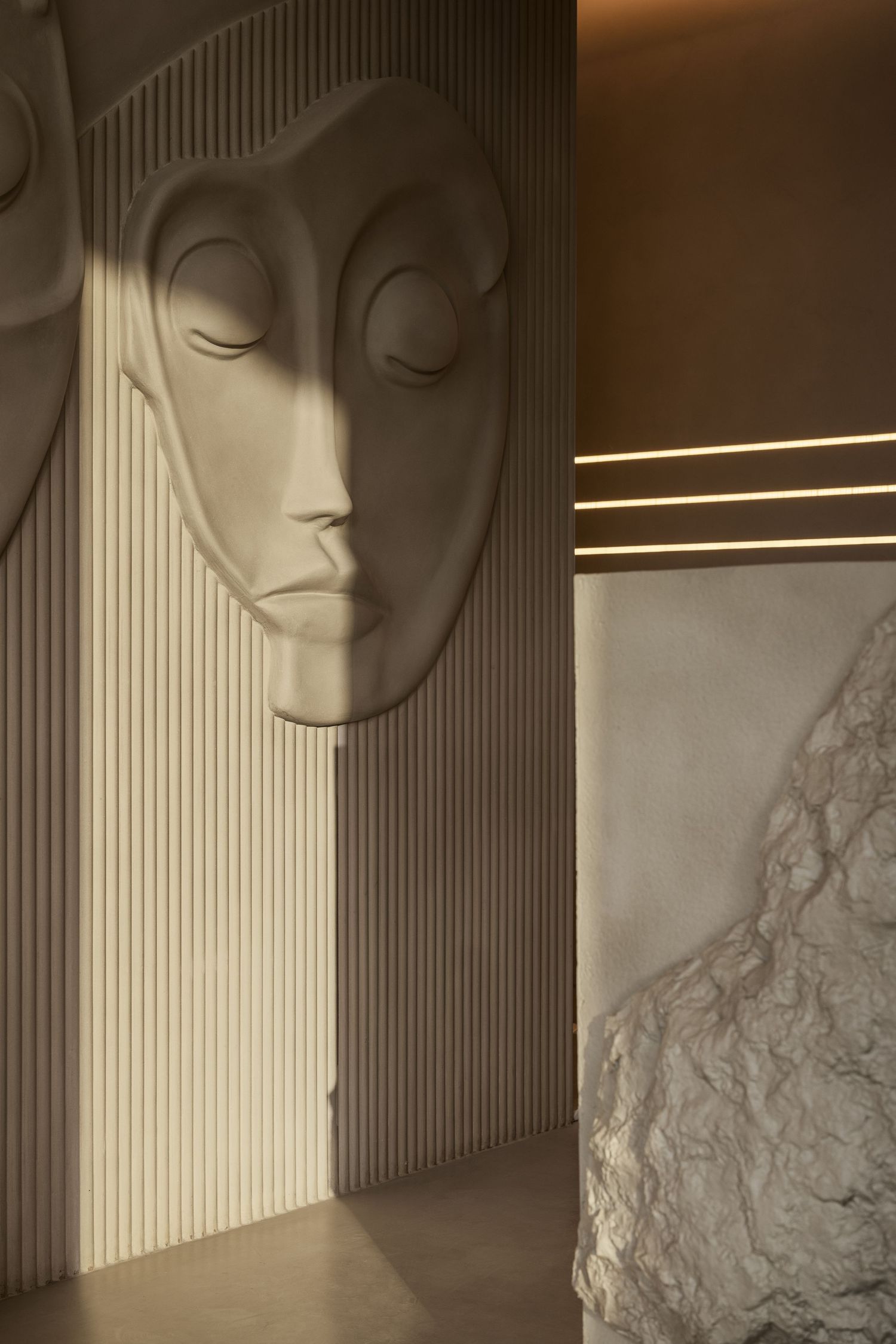Beige sculptural face relief on ribbed wall panel with warm LED strip lighting and rough stone element.