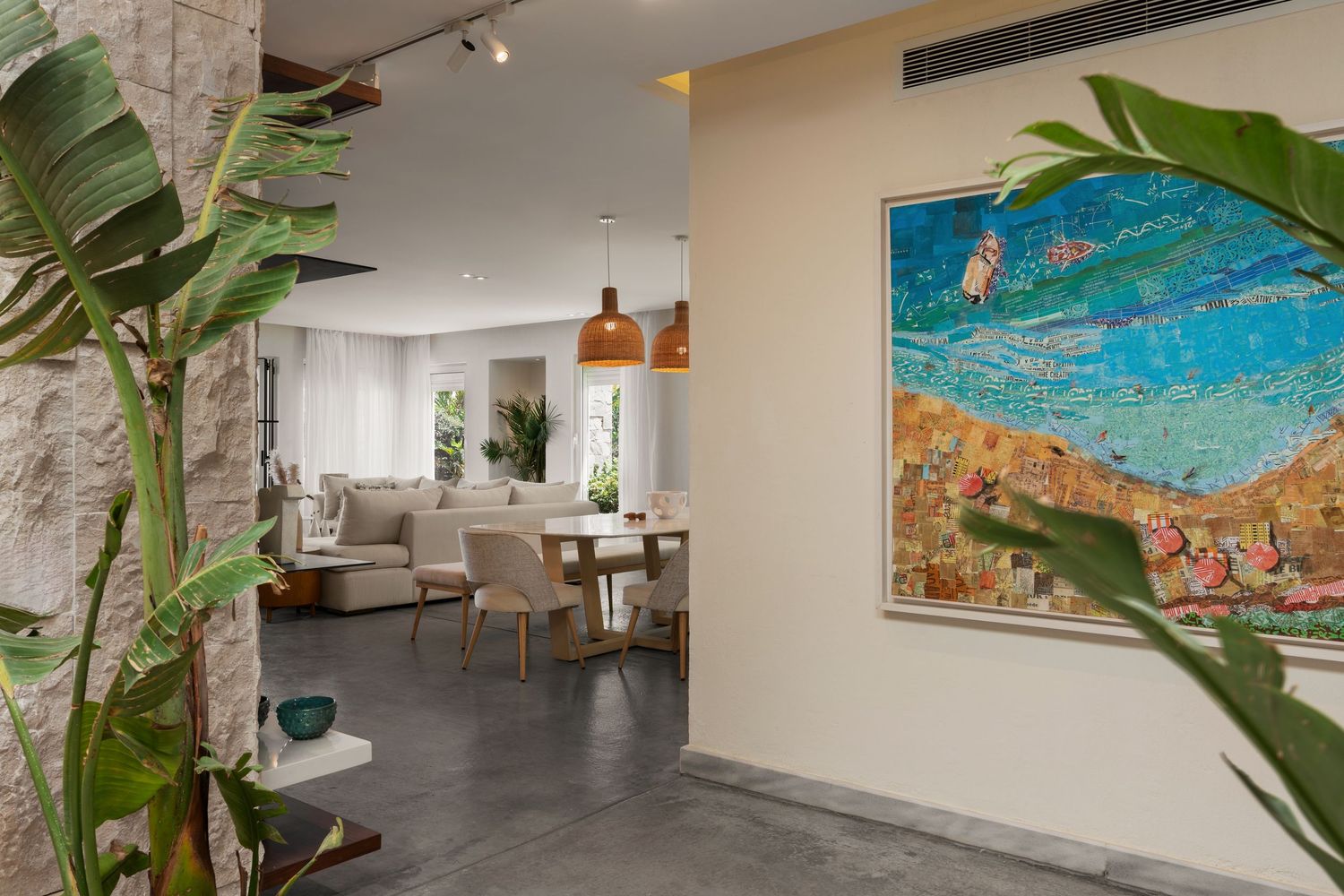 Stylish open-plan interior with tropical plants, large colorful artwork, dining area, and neutral toned furnishings.