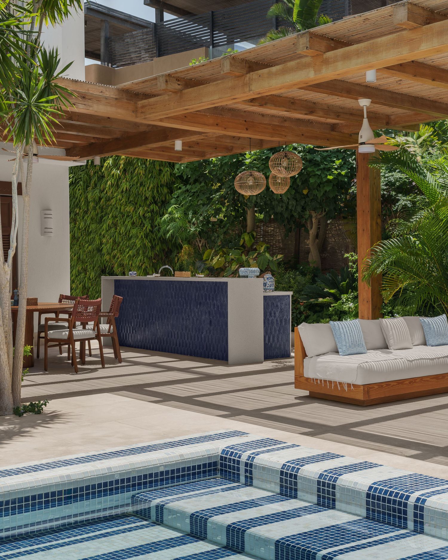 Luxury outdoor patio with wooden pergola, blue tiled pool, outdoor kitchen, and lounge seating surrounded by lush greenery.