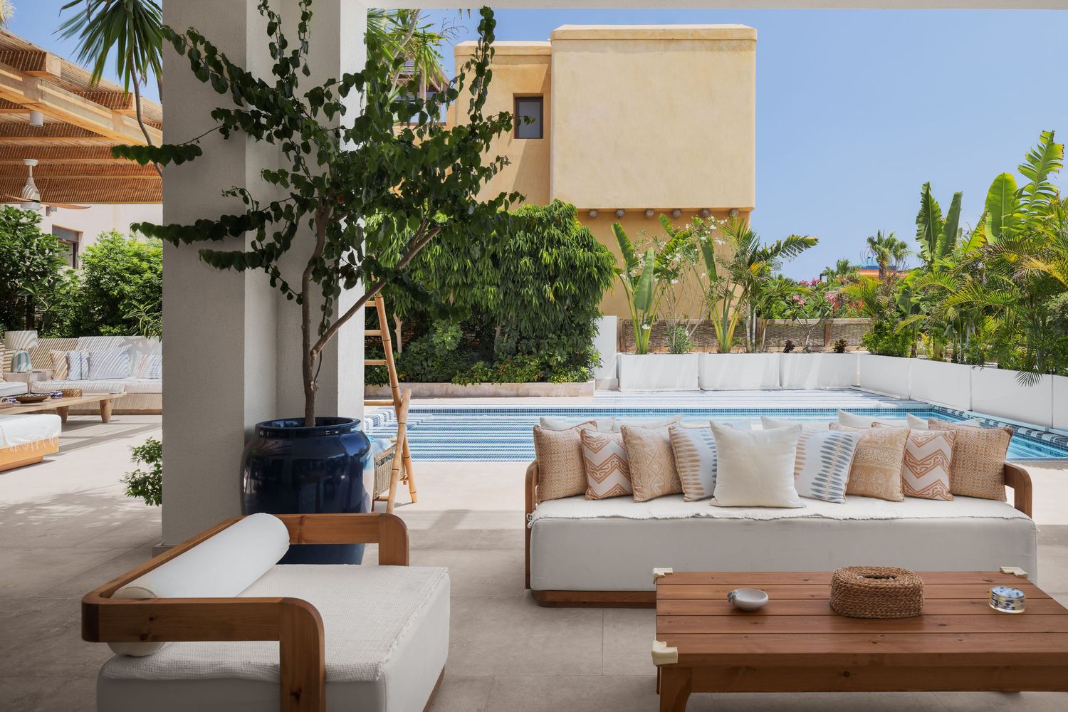 Bright outdoor living space with white upholstered sofas, wooden accents, blue ceramic pot, and pool view.