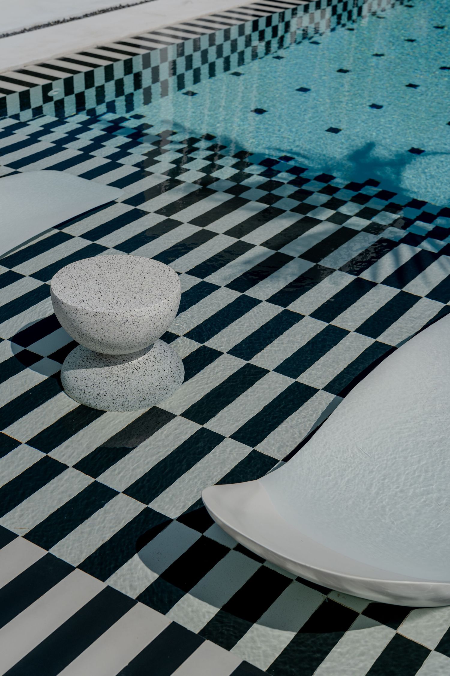 Black and white checkered pool deck with a sculptural white terrazzo stool beside a crystal blue swimming pool.