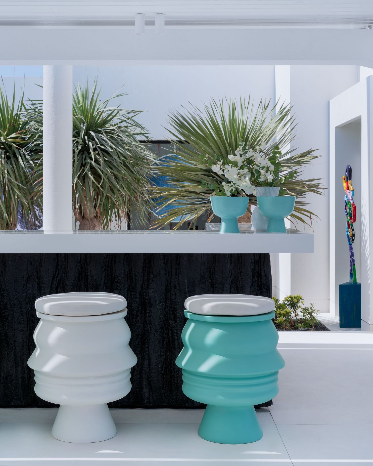 White and turquoise sculptural stools beside potted tropical plants on a sleek white outdoor terrace.