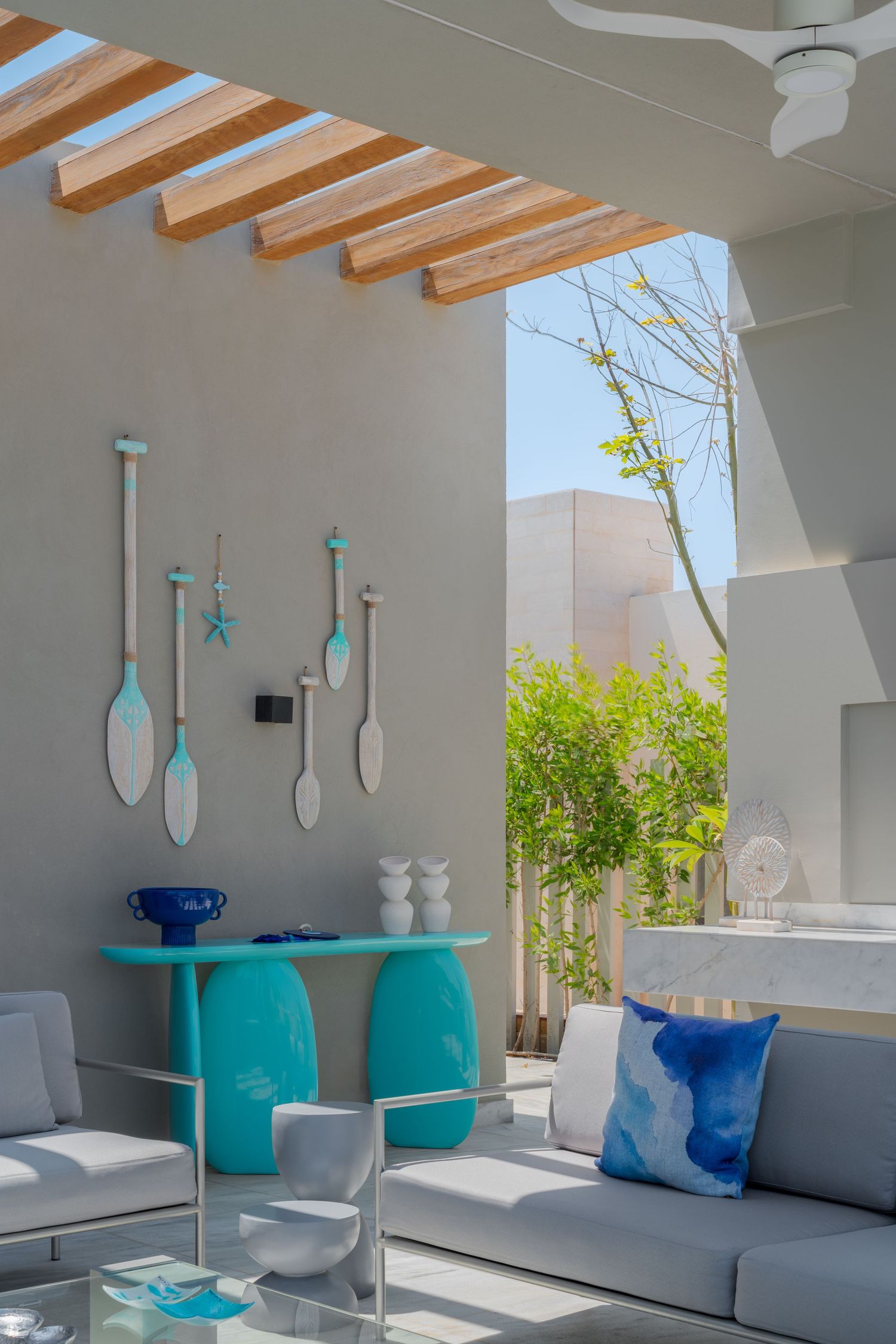 Stylish outdoor patio with turquoise console table, decorative oar wall art, and gray sofas under wooden pergola.