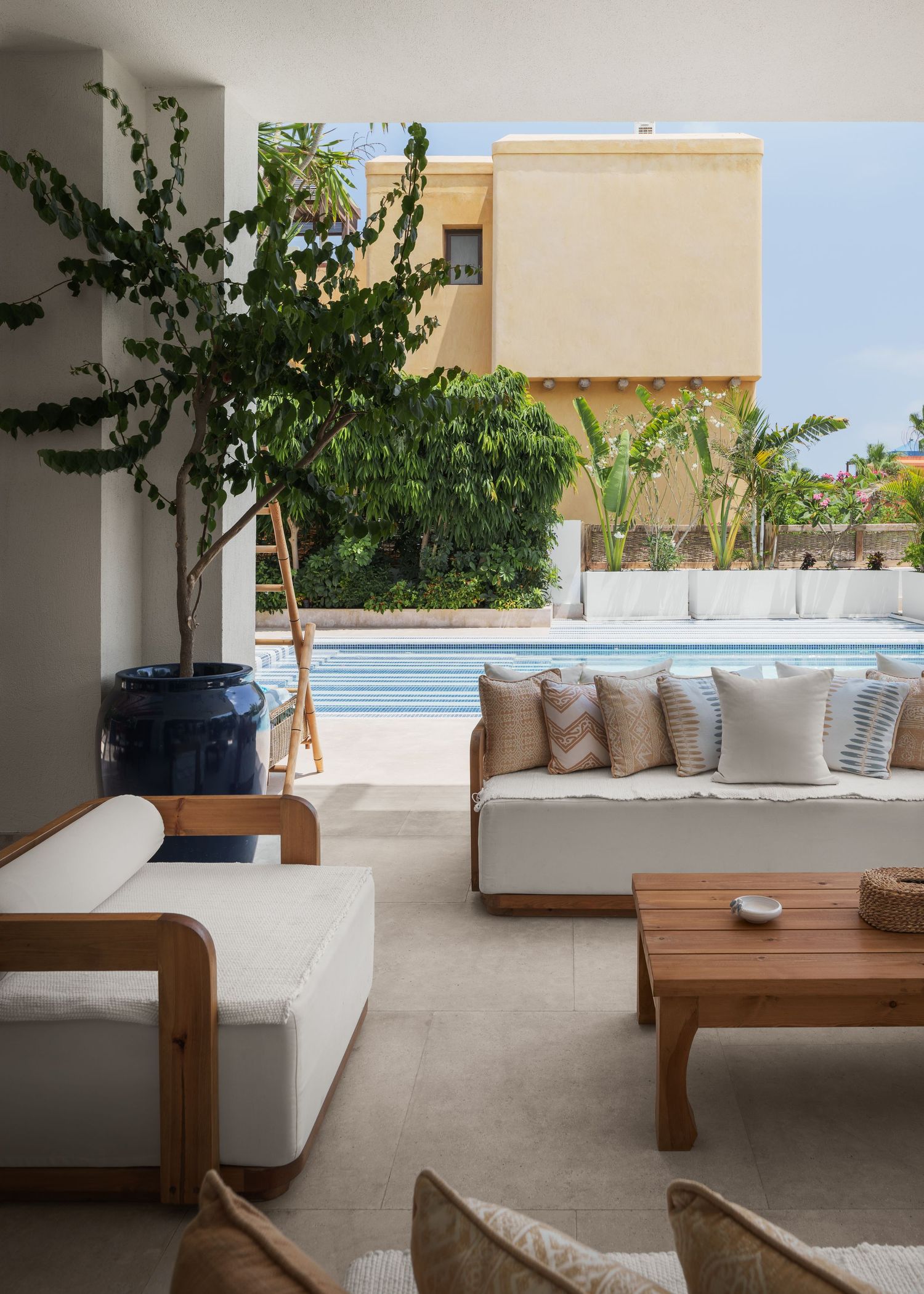 Modern covered terrace with white sofas, wooden coffee table, large potted tree, and swimming pool view.