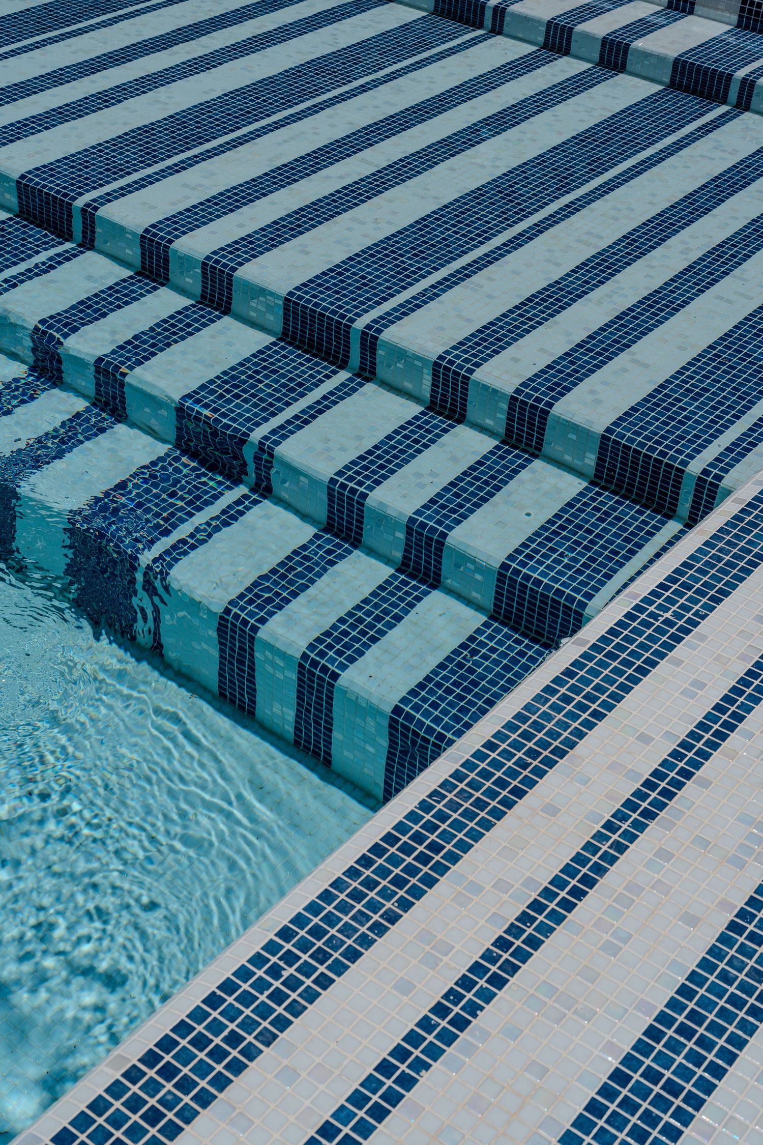Geometric blue and white mosaic tile pool steps with striped pattern submerged in clear turquoise water.