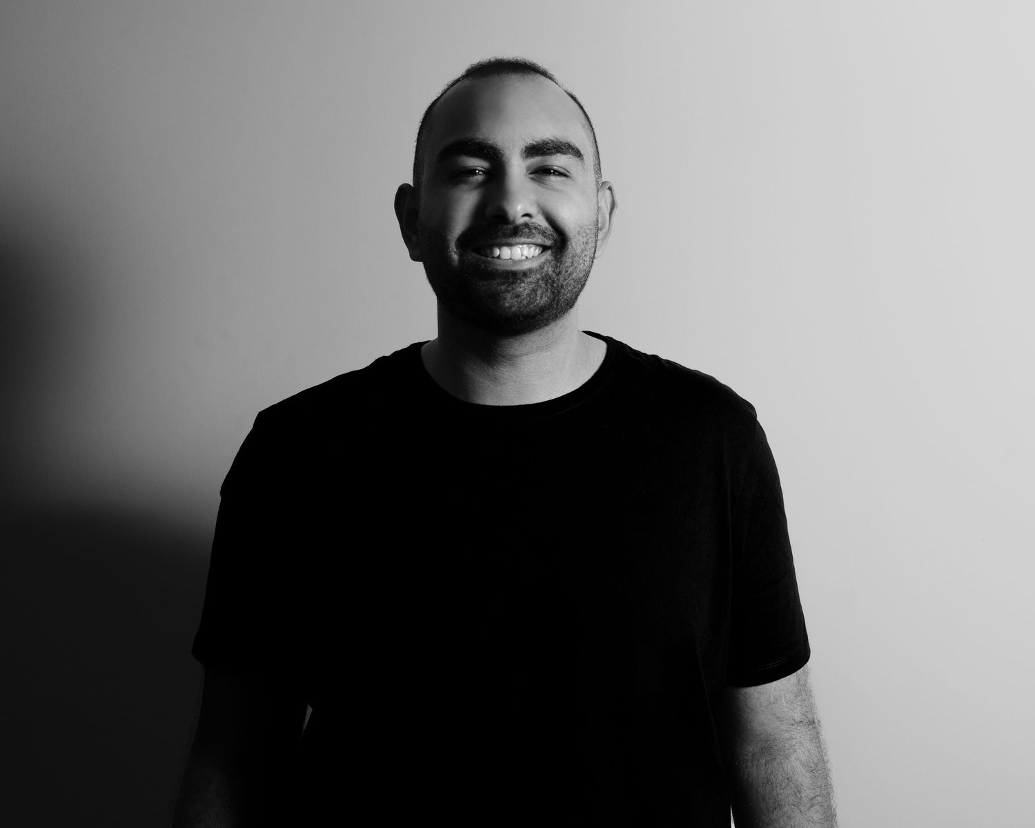 Black and white portrait of a smiling bearded man in a black t-shirt against a neutral gray background.