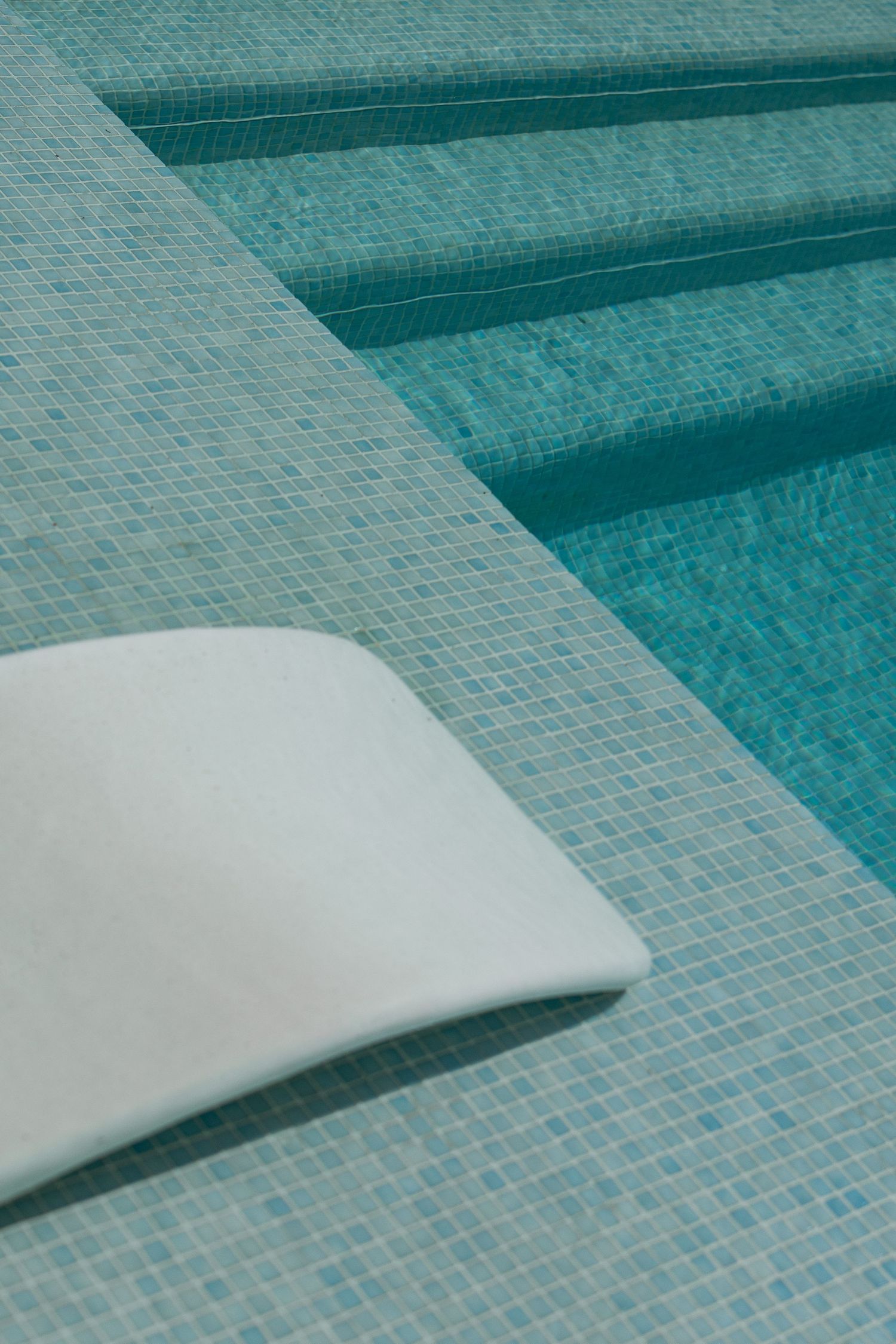 Close-up aerial view of a white lounge chair beside a turquoise mosaic tile pool with shallow entry steps.