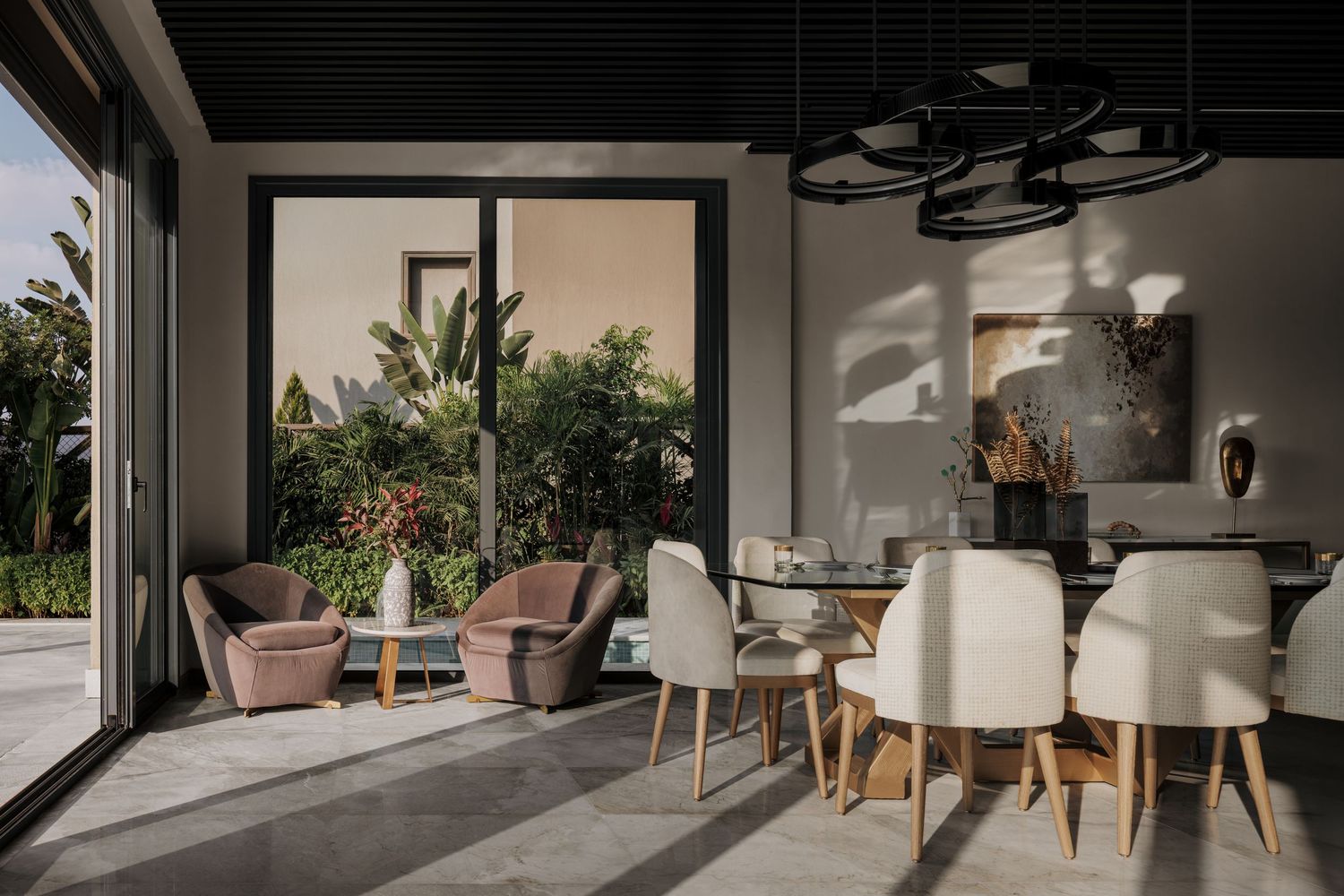 Modern open-plan dining area with white upholstered chairs, velvet accent seating, large windows, and lush tropical garden views.