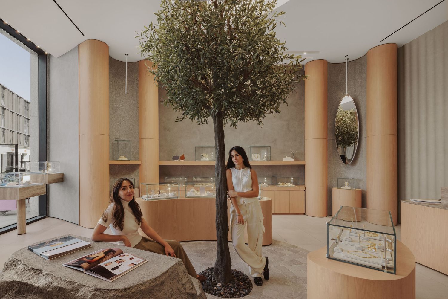 Two women pose in a luxury jewelry boutique featuring a live olive tree, stone seating, and warm wood display cases.