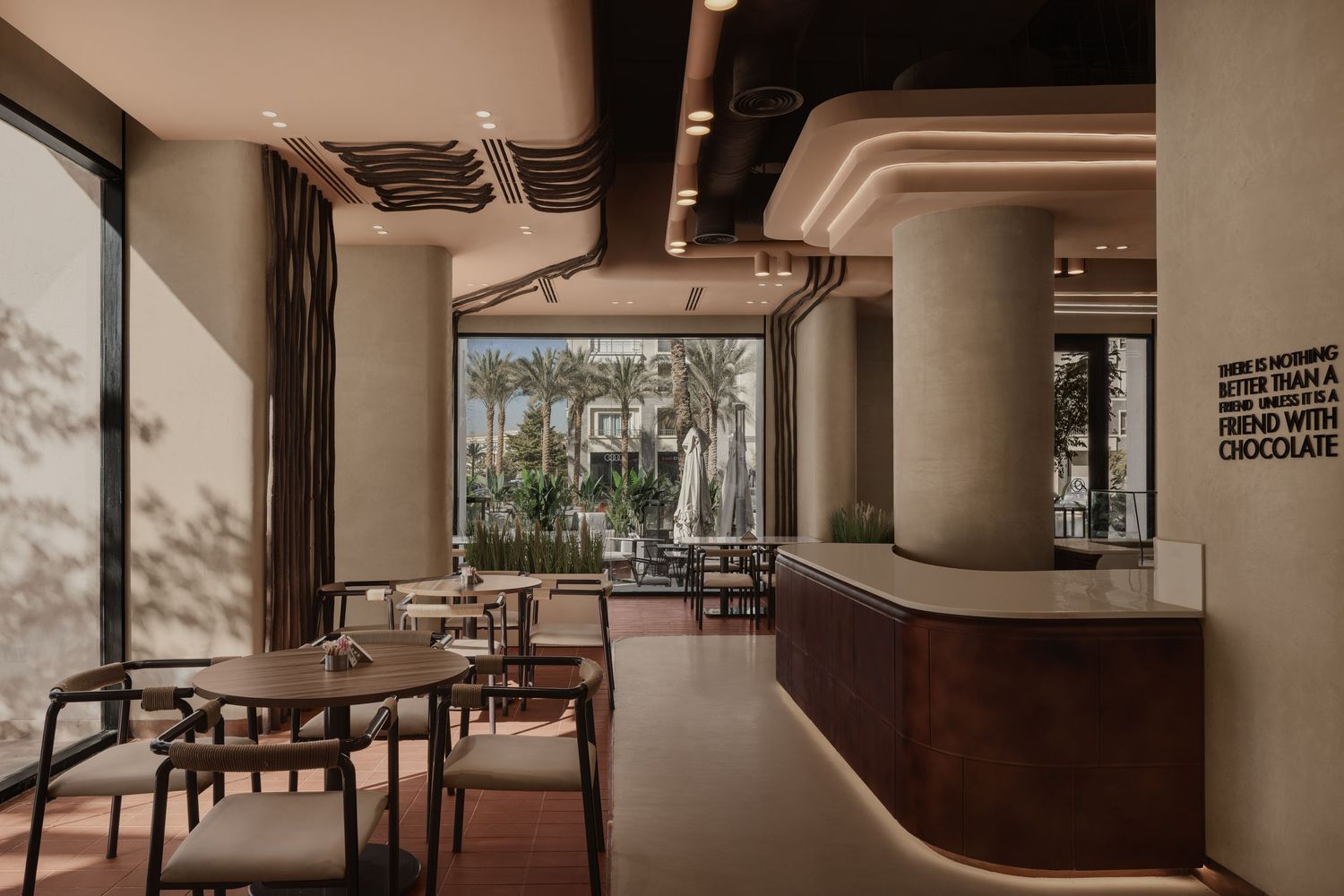 Modern cafe interior with dark wood furniture, curved reception desk, and large windows overlooking palm trees.