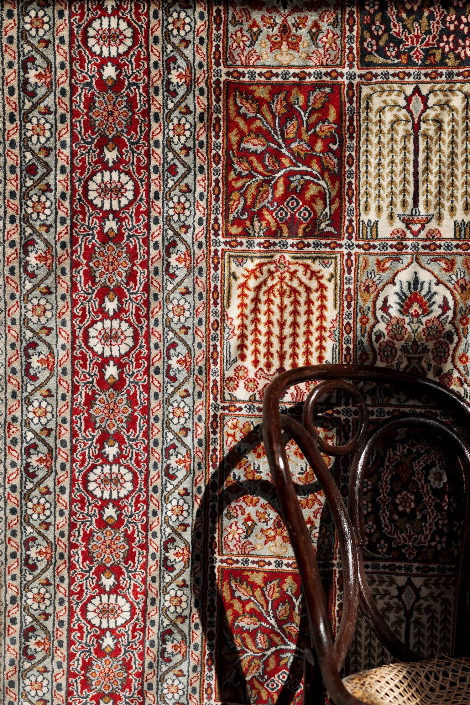 Ornate traditional carpet with red floral patterns displayed on wall beside a vintage bentwood chair.