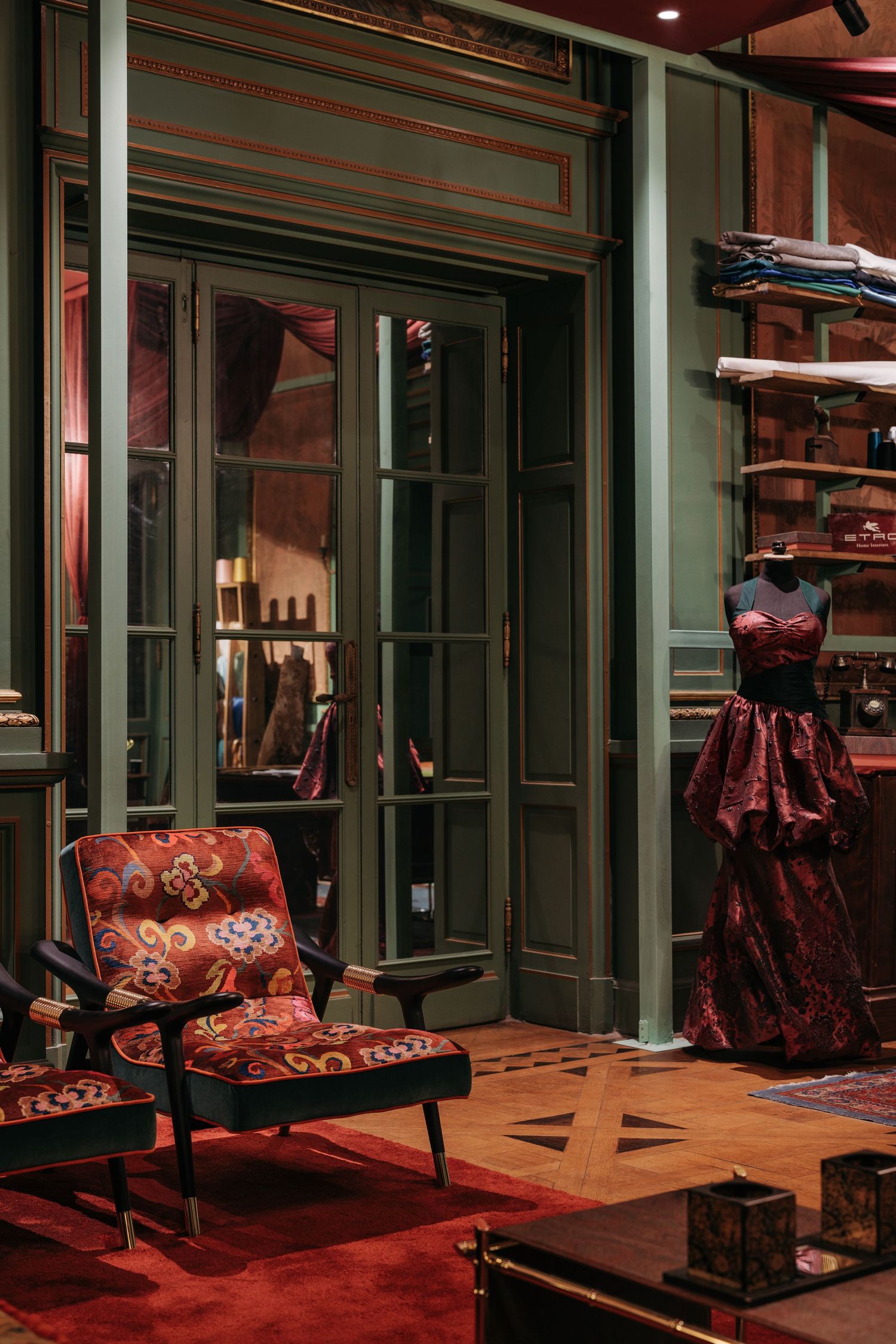 Elegant dark green boutique interior with red gown on mannequin, patterned armchair, and French doors.