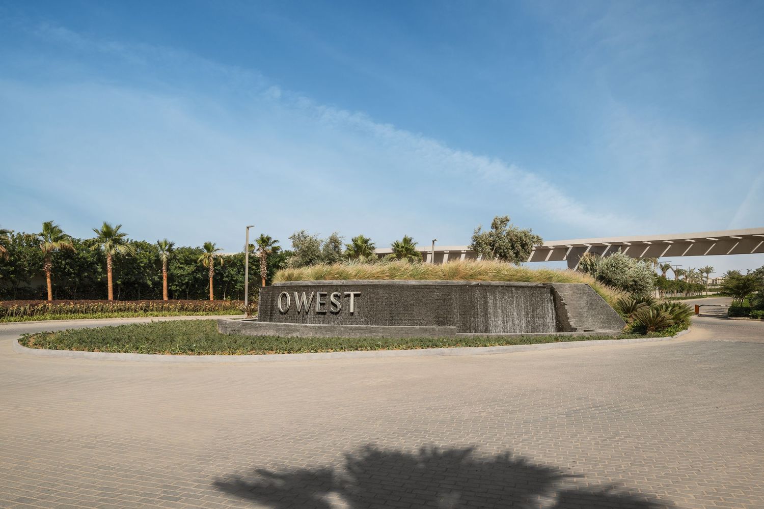 Panoramic view of OWEST community entrance roundabout with palm trees, landscaping, and iconic gateway structure.