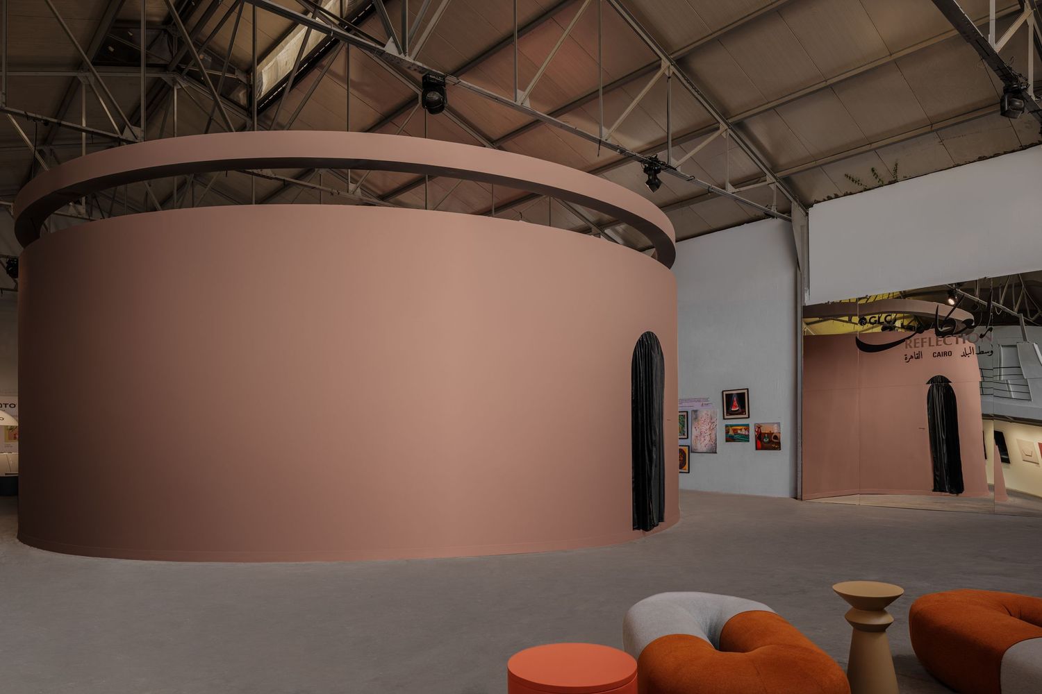 Large dusty pink cylindrical structure with black arched doors displayed in spacious industrial art fair hall.