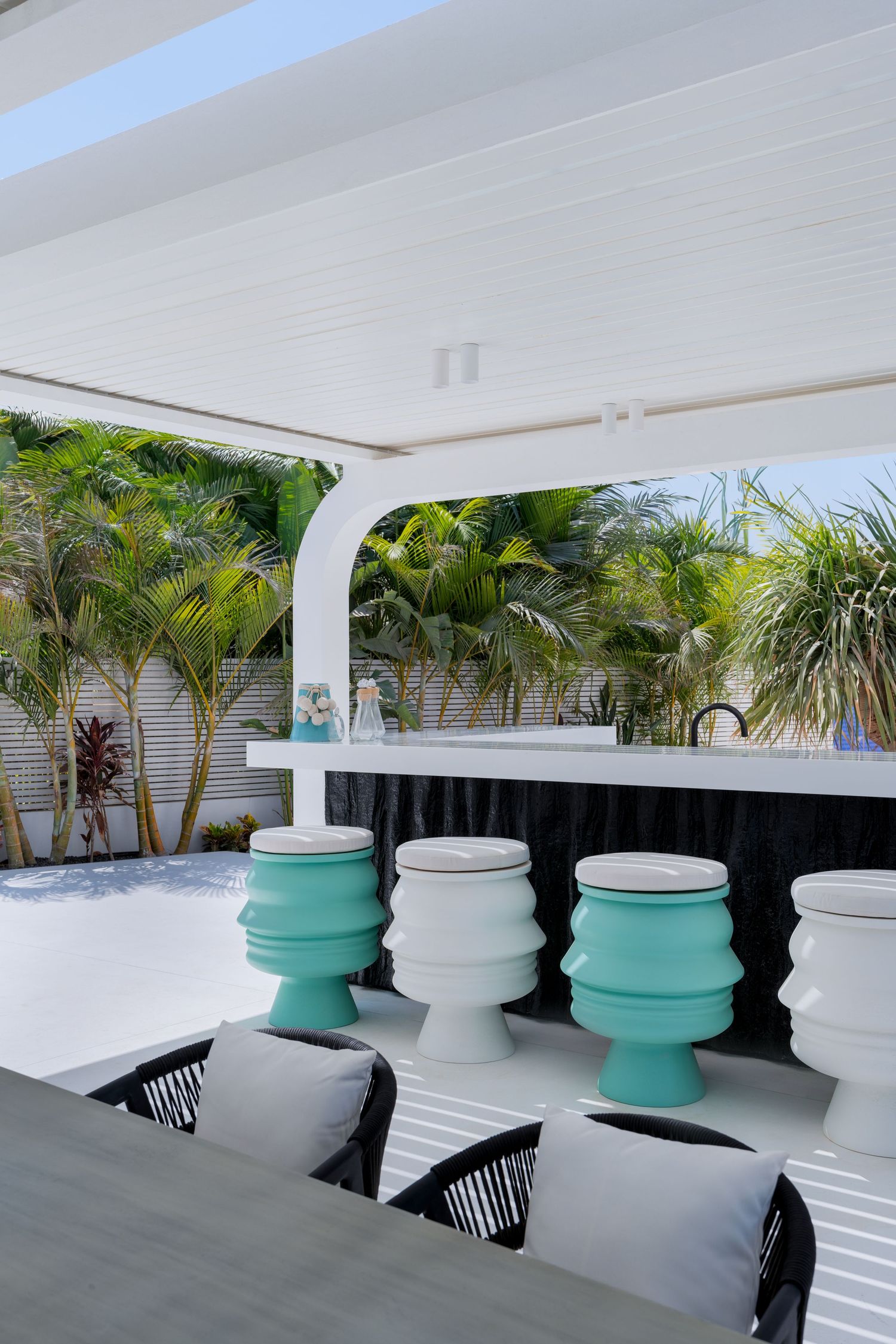 Modern outdoor bar with teal and white sculptural stools under a white pergola surrounded by tropical palms.