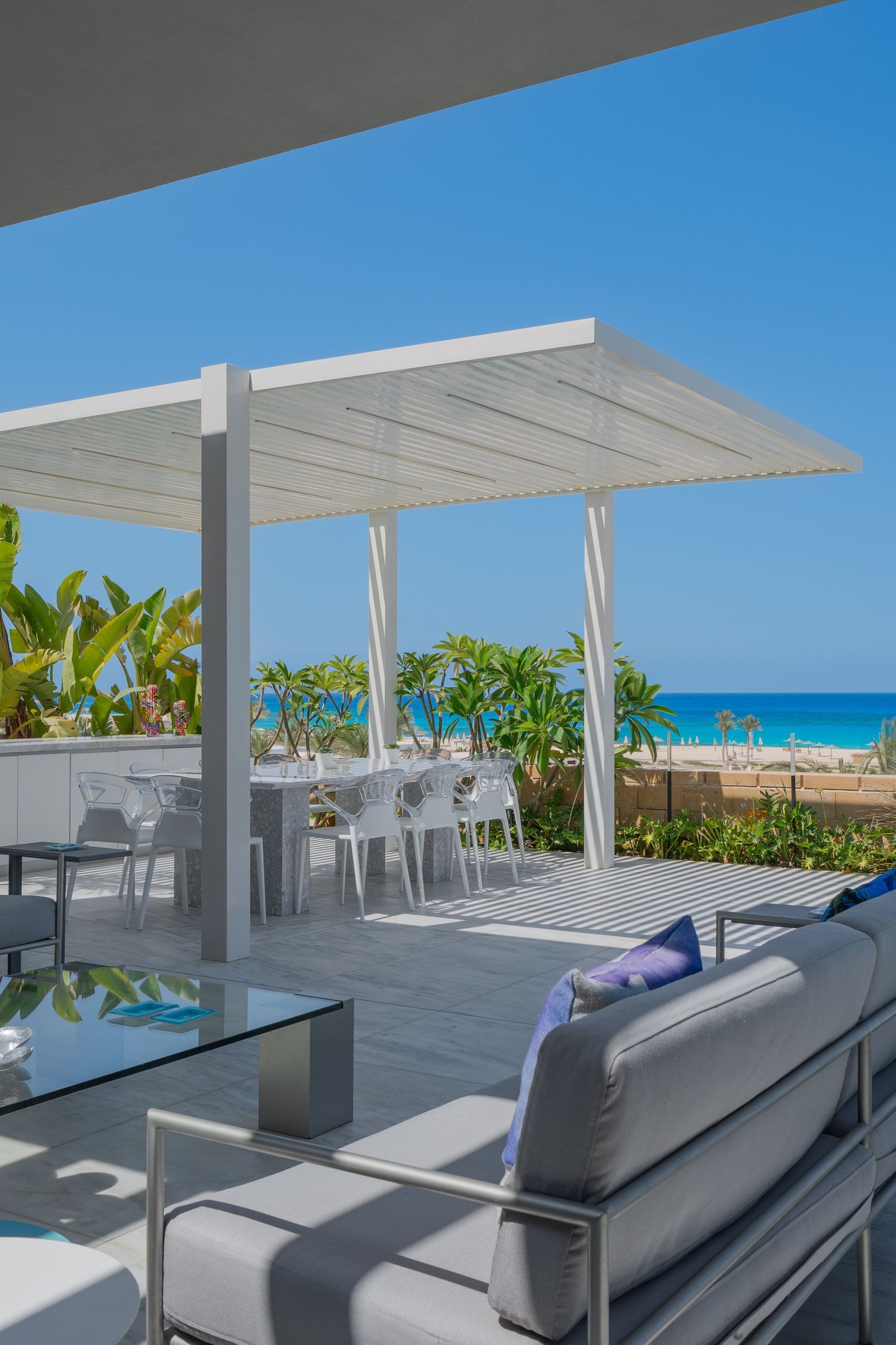 Elegant beachfront terrace with white pergola, outdoor dining set, gray sofa, and stunning ocean view.