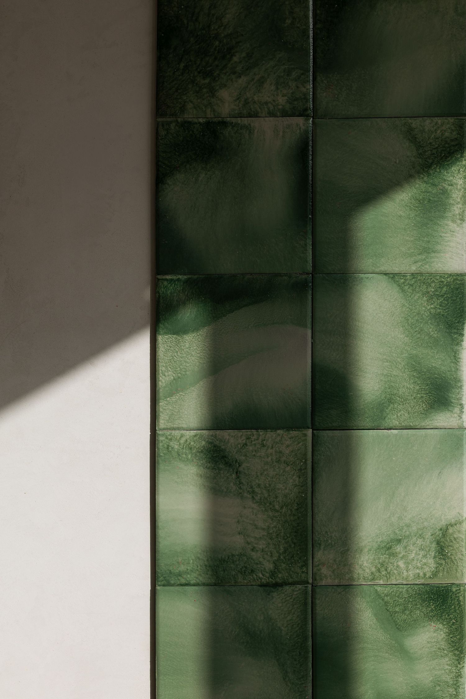 Deep green glossy ceramic wall tiles with light and shadow patterns next to white plaster wall.