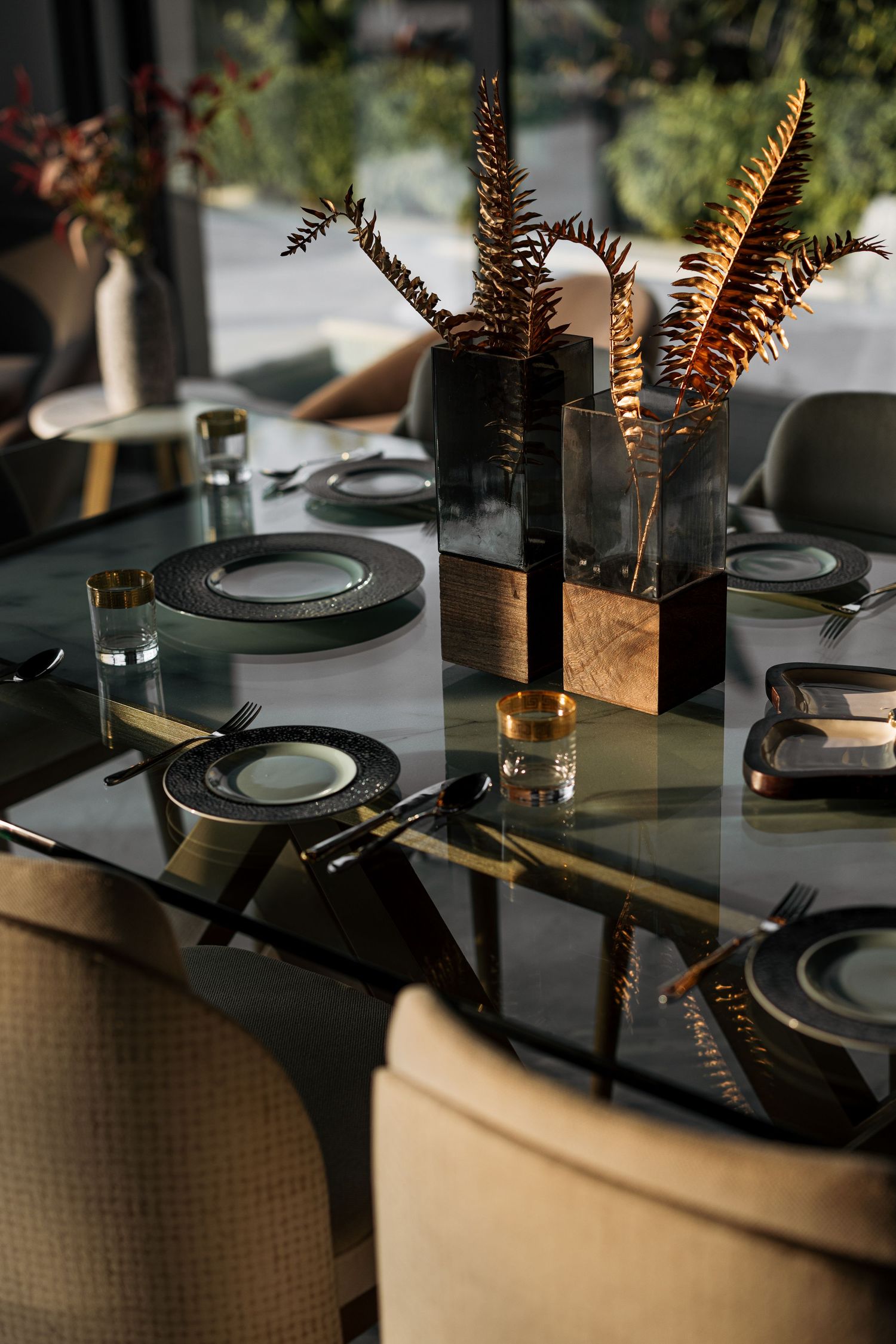 Elegant glass dining table set with dark ceramic plates, gold accents, and bronze fern centerpieces against a garden backdrop.