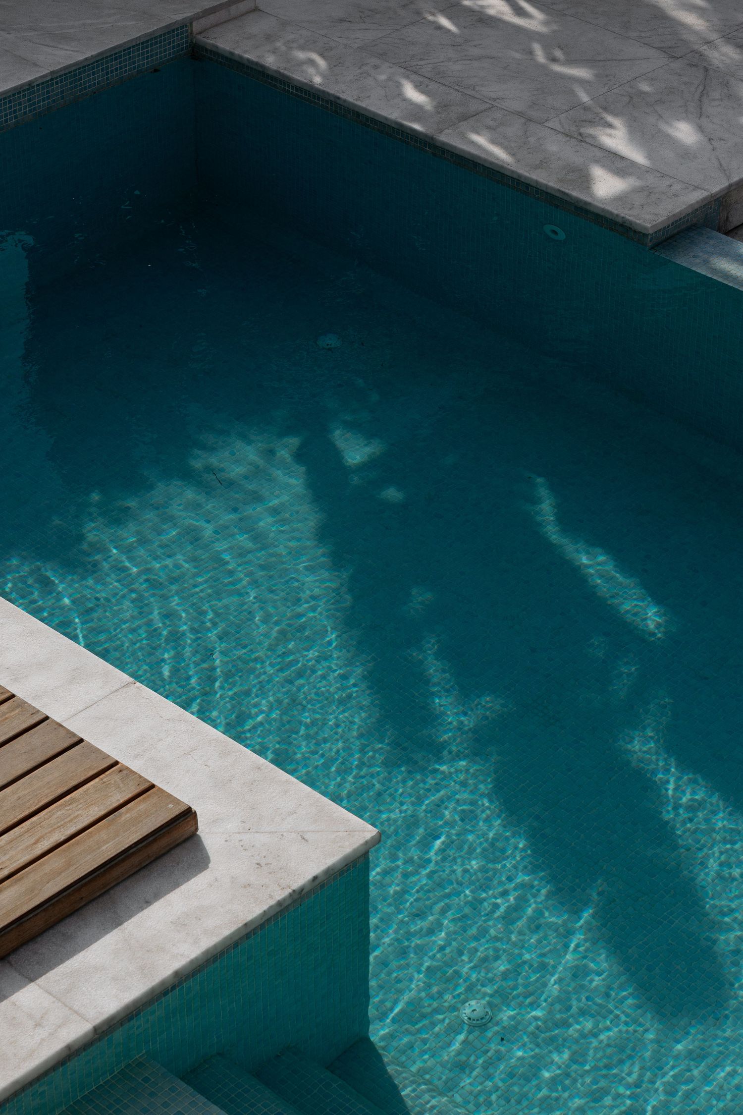 Aerial view of turquoise swimming pool with wooden deck and marble surround casting shadows.