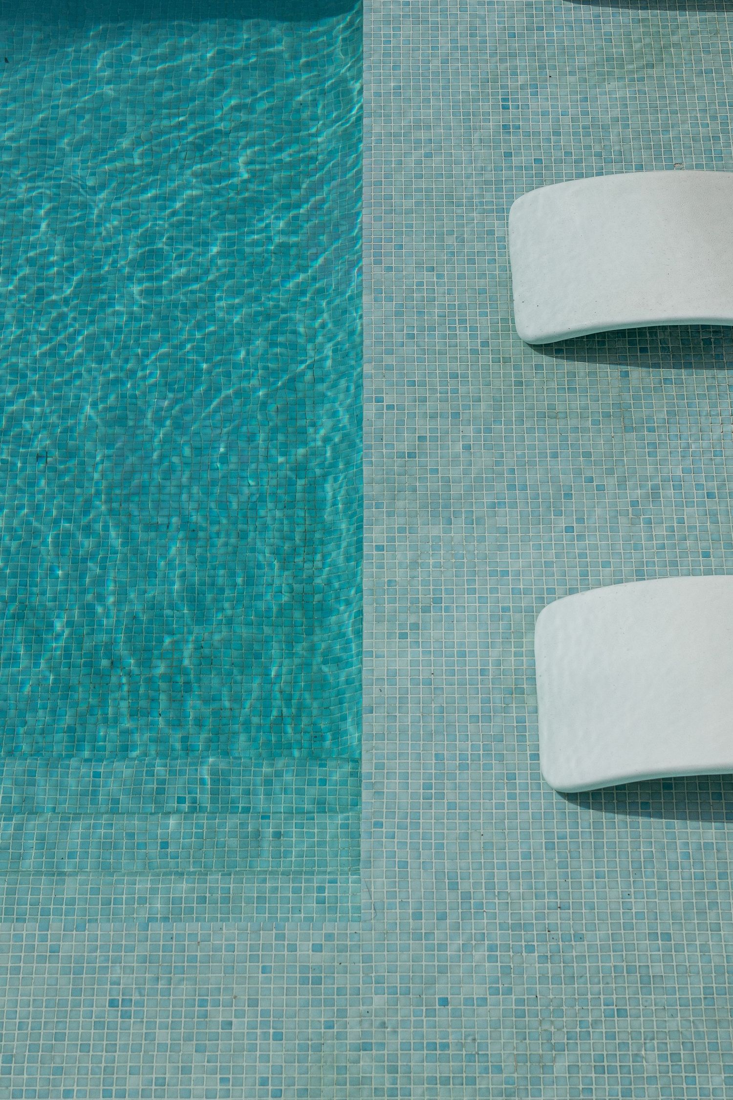 Overhead view of a turquoise swimming pool with two white lounge chairs on mosaic tile poolside.