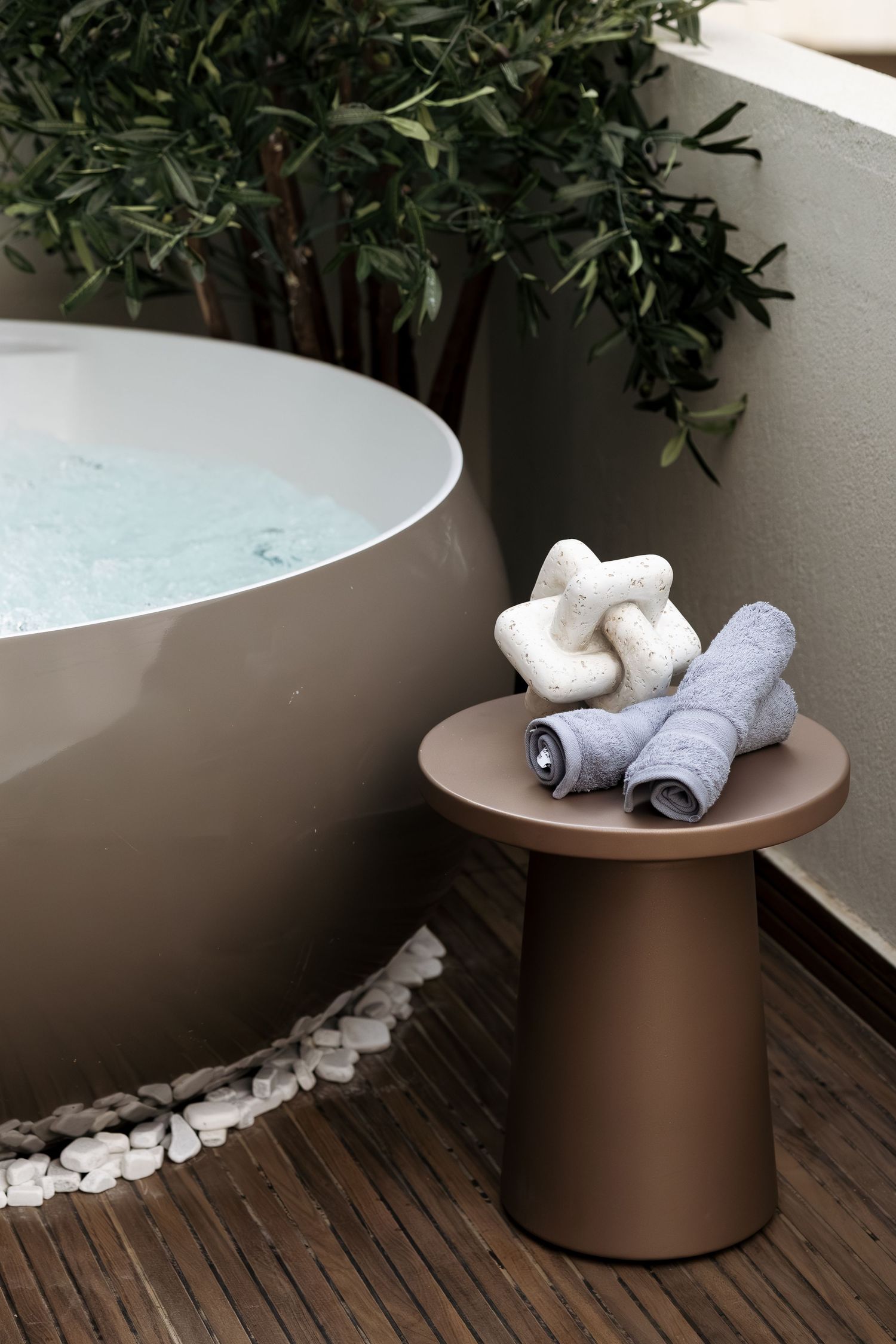 Outdoor spa area featuring a round freestanding bathtub with pebble surround, bronze side table, and rolled towels.