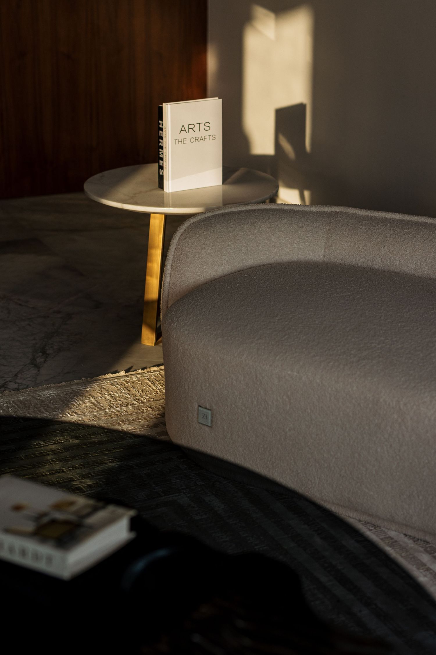 Close-up of curved gray upholstered sofa beside gold side table with arts book in warm sunlight interior.