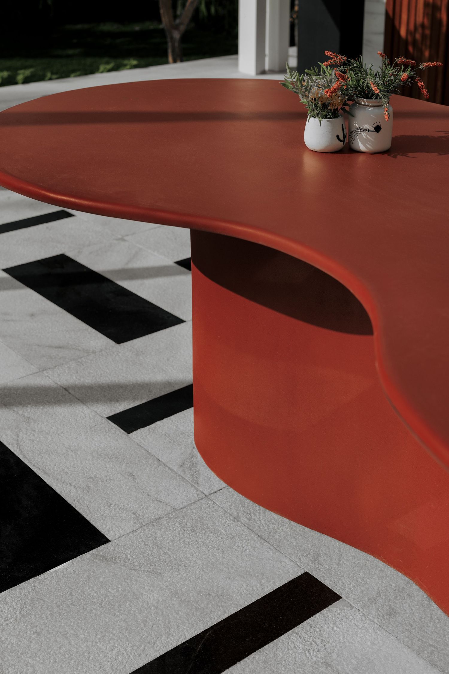 Close-up of sculptural red outdoor dining table with organic curved base on geometric black and white tiles.