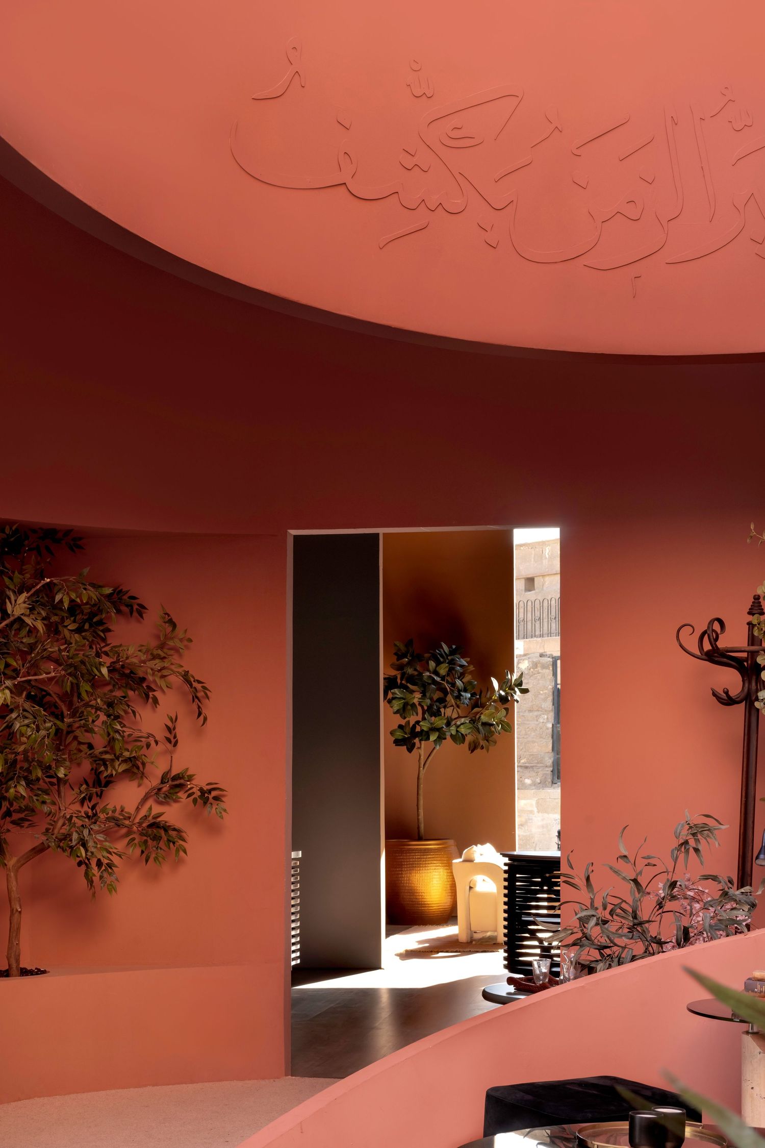 Pink curved interior with embossed Arabic ceiling script, olive trees, and arched doorway.