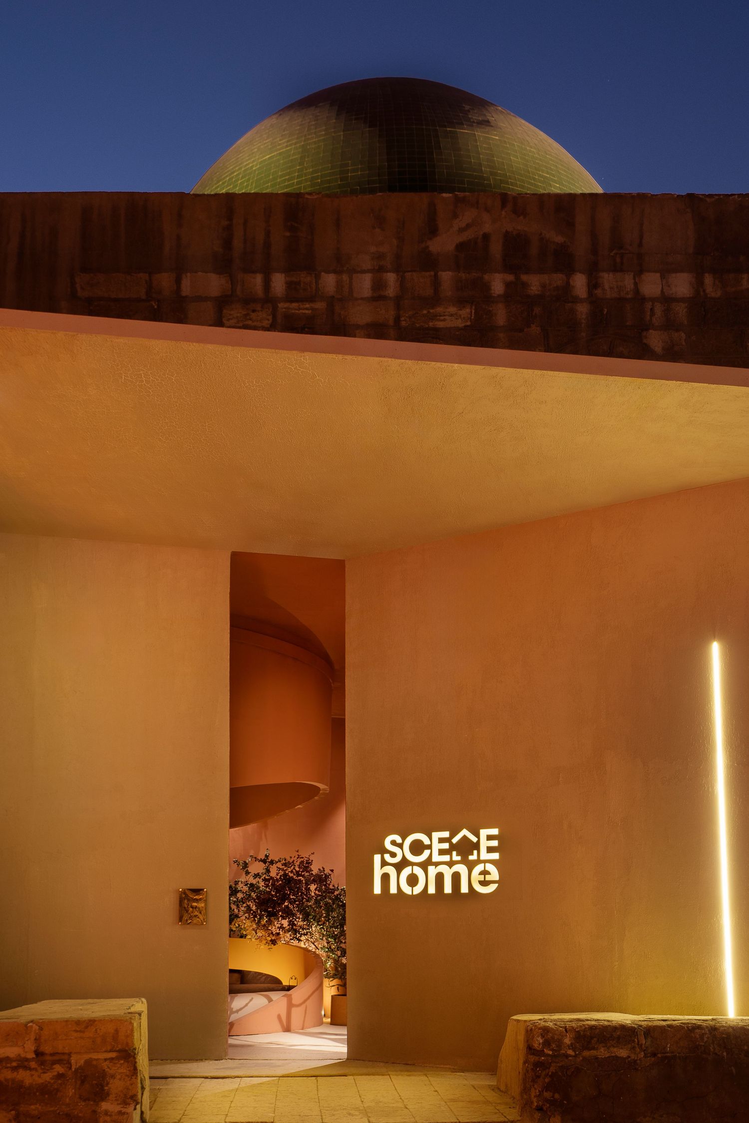 SCECE Home storefront at dusk with illuminated signage, curved architecture, and green dome against blue sky.