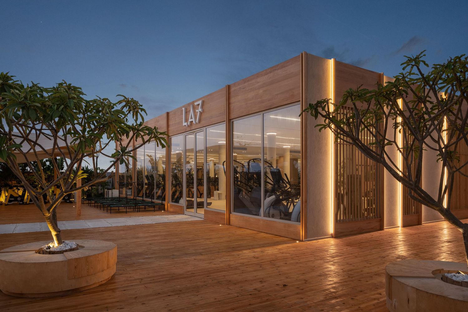 Modern LA7 gym building with floor-to-ceiling glass walls, warm wood accents, and tropical frangipani trees illuminated at dusk.