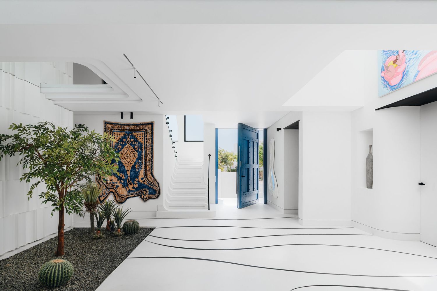 Modern white interior with a Persian rug, cactus garden, blue door, and abstract art in a bright open-plan space.