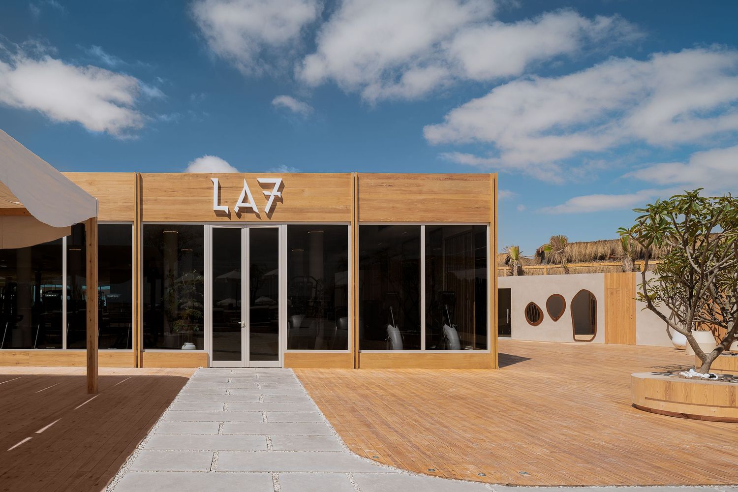 Modern LA7 building exterior with warm wood cladding, large black windows, and decorative frangipani tree at entrance.