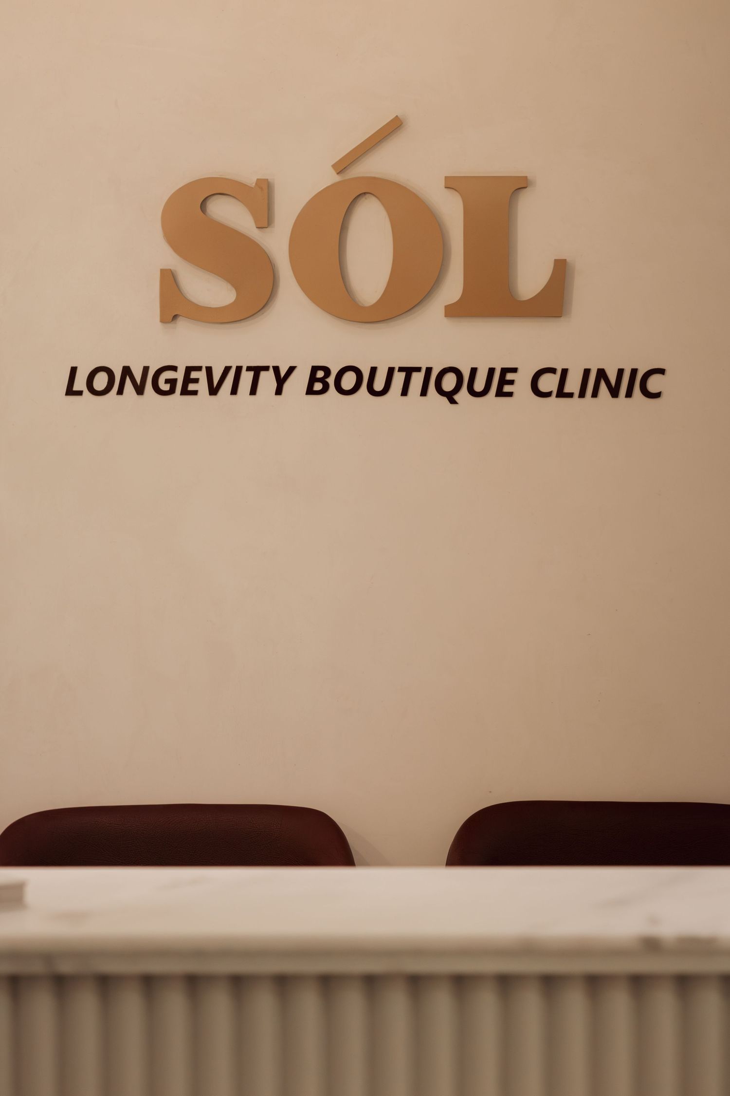 Close-up of SOL Longevity Boutique Clinic gold lettering sign mounted on beige wall above reception desk.