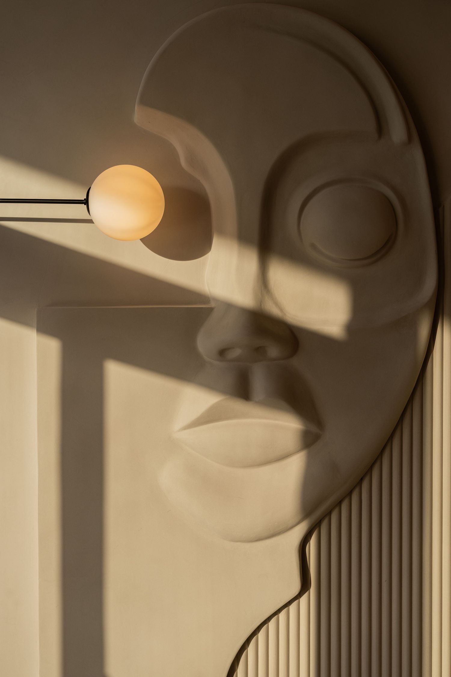 Abstract plaster face wall sculpture with globe lamp casting dramatic shadows on beige wall.