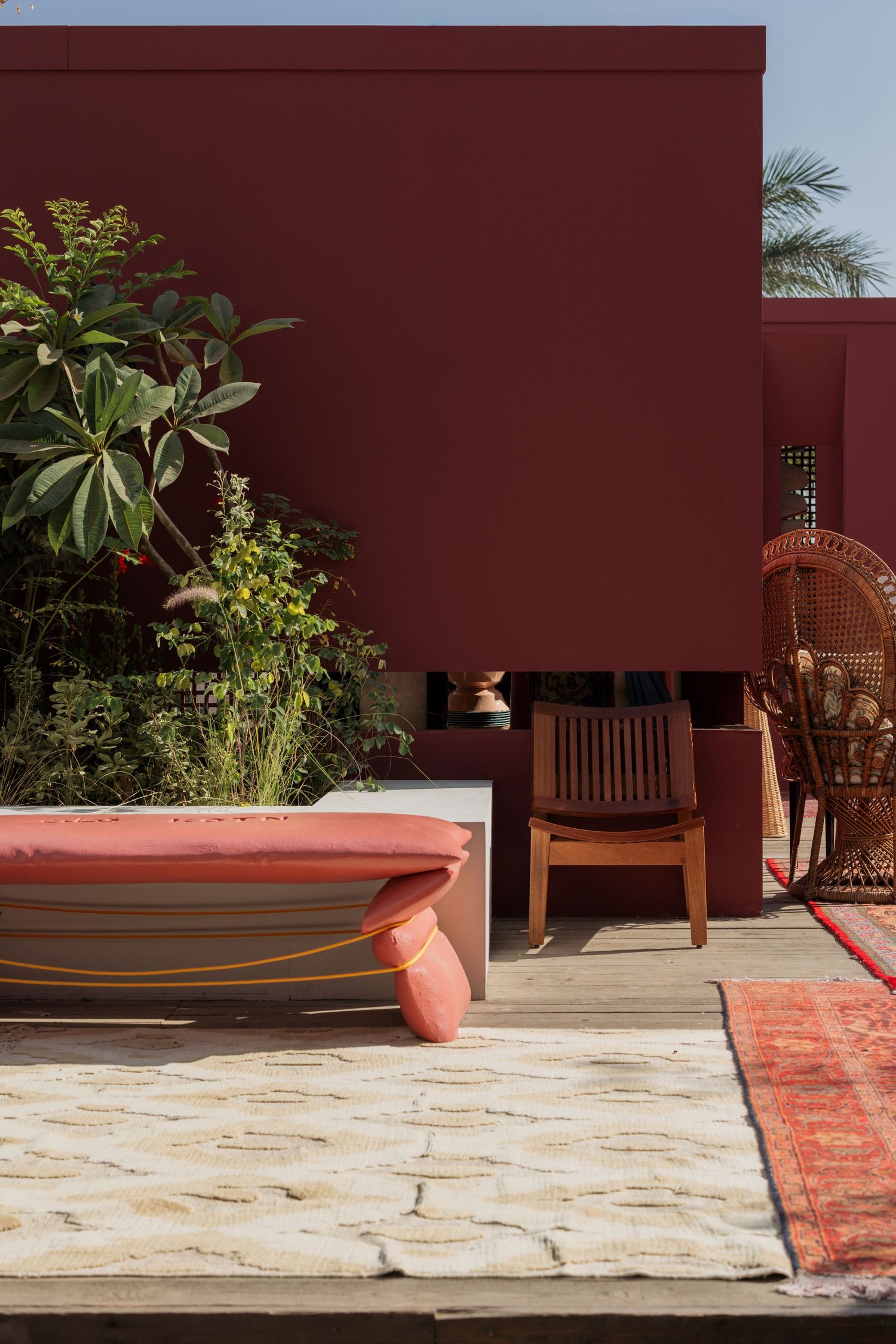 Pink cushioned outdoor daybed beside wooden chair on terrace with deep red wall and tropical greenery.