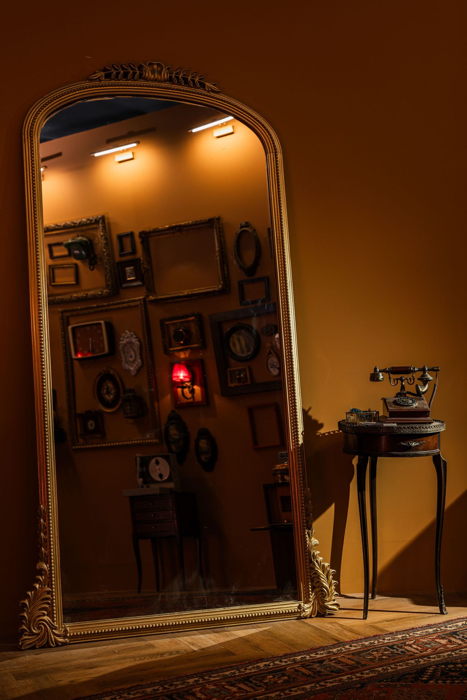 Large ornate gold mirror reflecting a wall of vintage frames and an antique telephone on a side table.