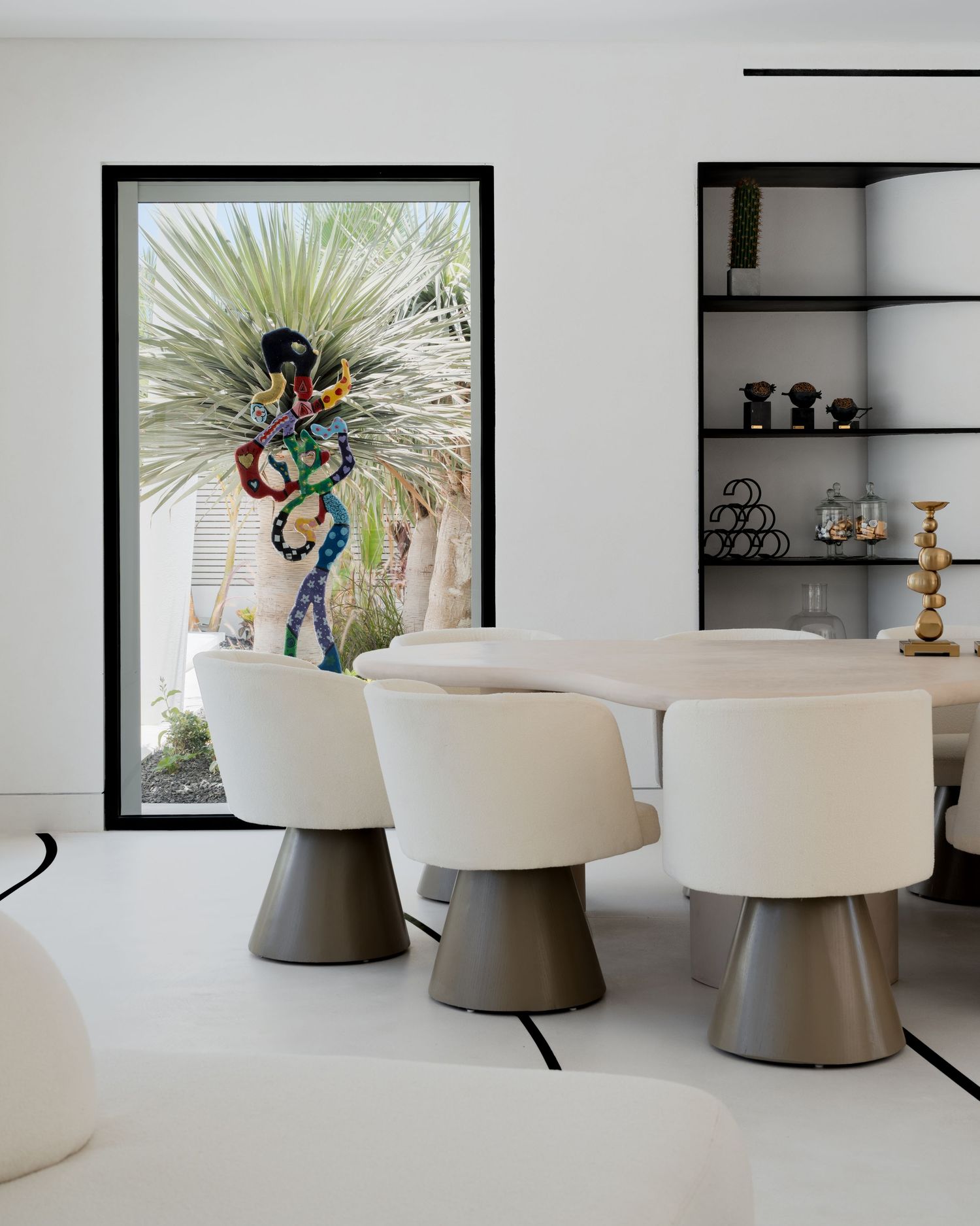 Minimalist white dining space with cream chairs, stone table, and framed tropical garden sculpture view.