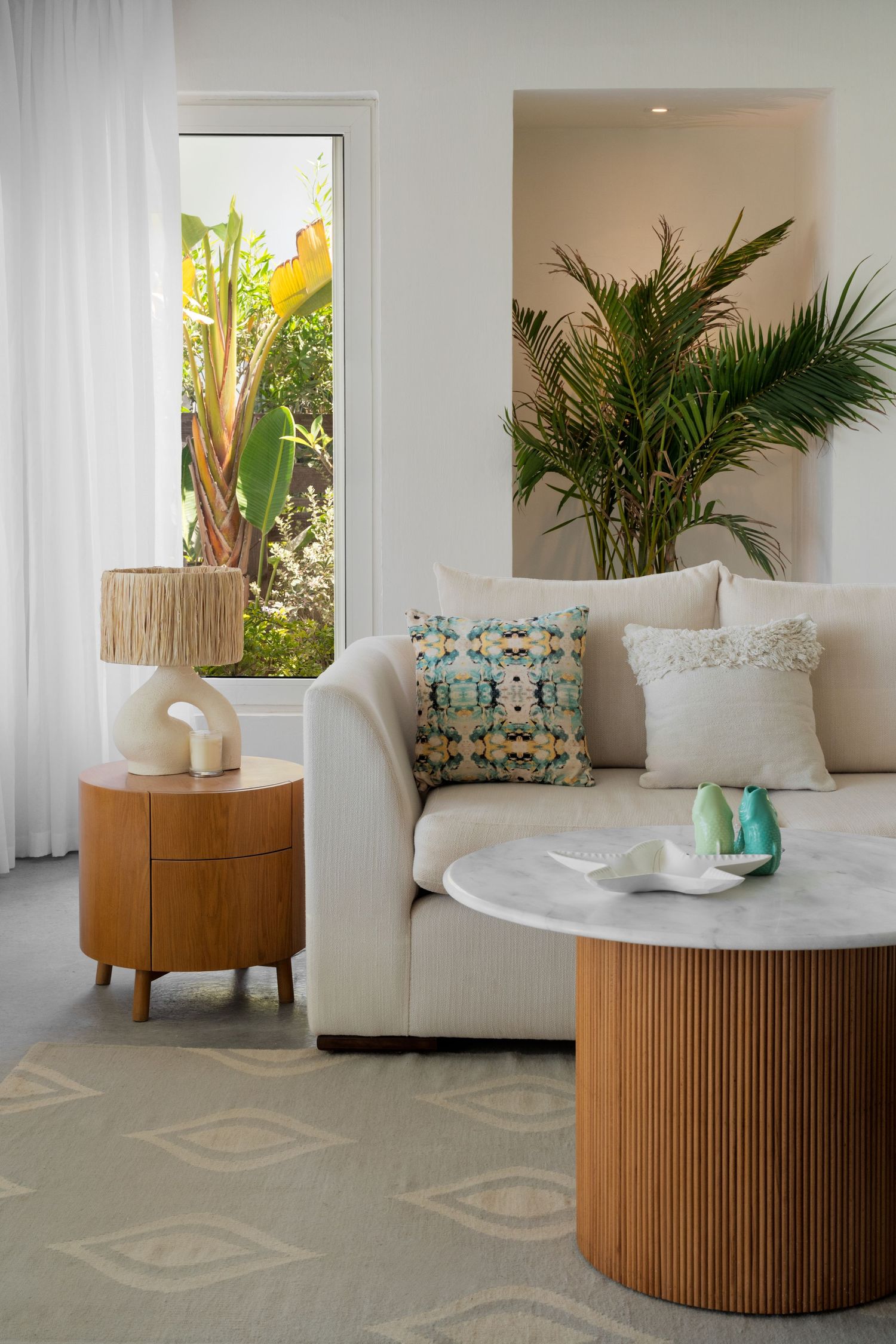 Cozy living space with beige sofa, patterned cushions, marble coffee table, and tropical plants by bright window.
