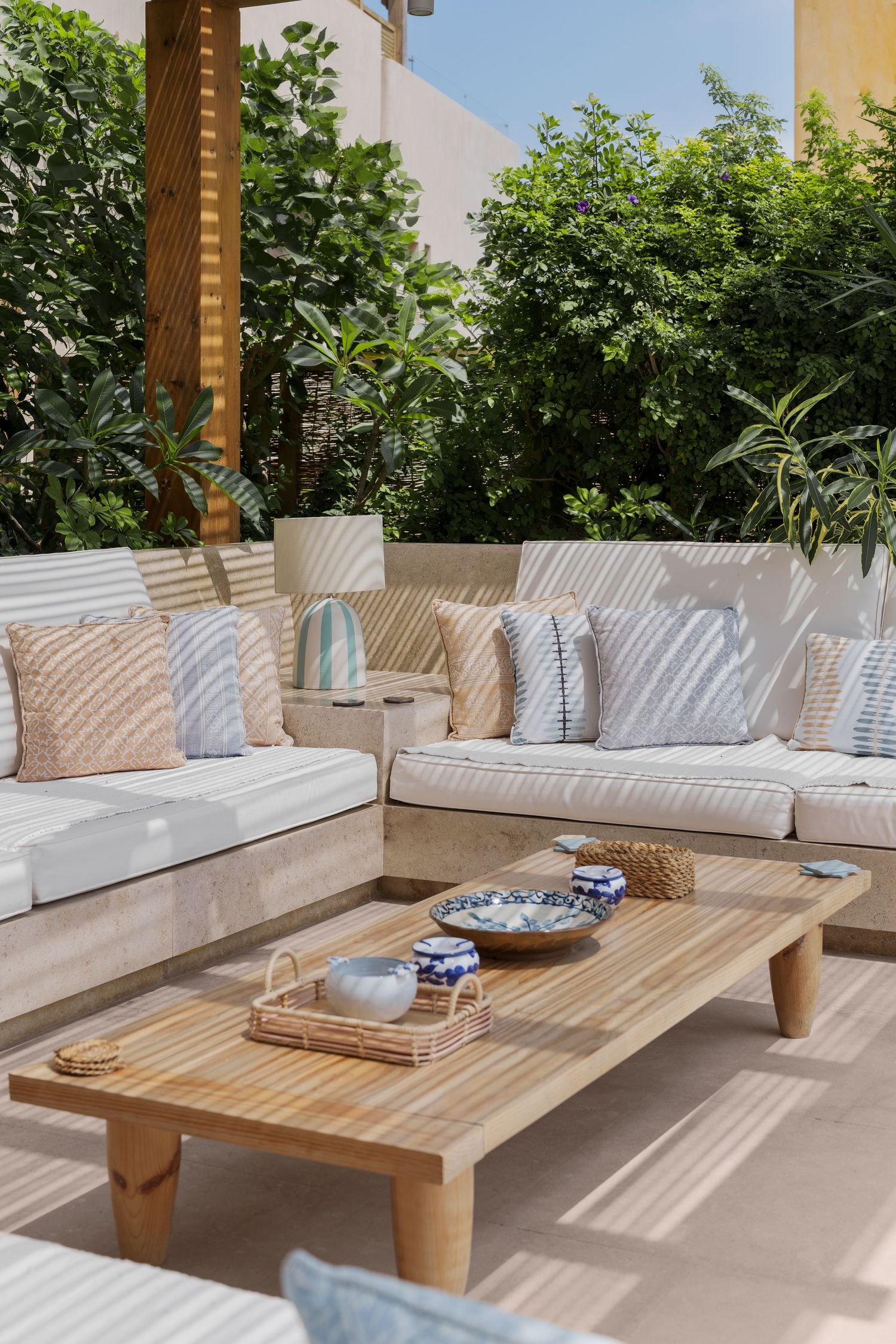 Outdoor lounge area with white cushioned sofas, striped pillows, and a wooden coffee table surrounded by tropical plants.