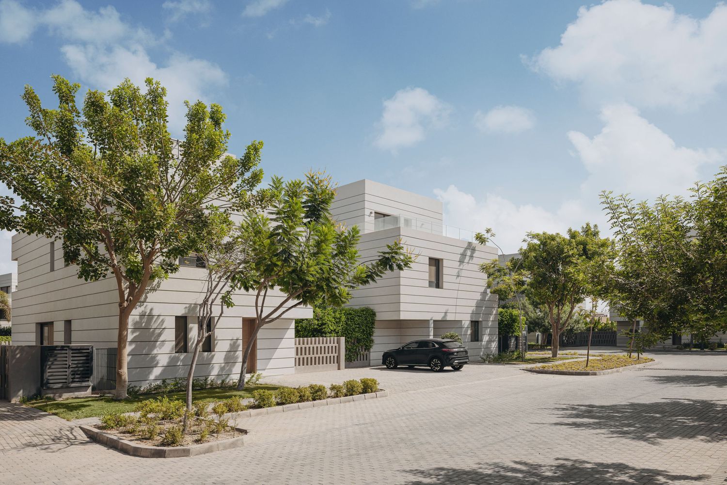 Contemporary white cubic villas surrounded by mature trees with a black car parked on paved driveway under bright sky.