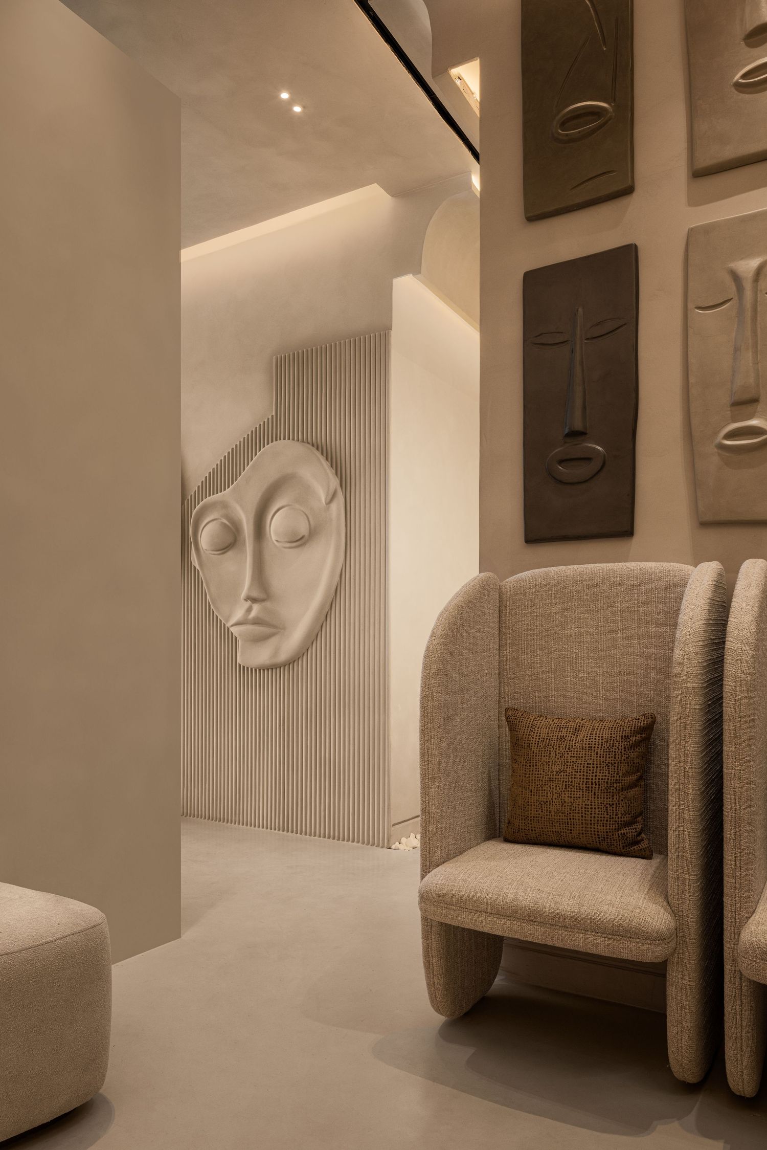 Minimalist beige spa lounge with sculptural face wall art, a cozy wingback chair, and soft ambient lighting.