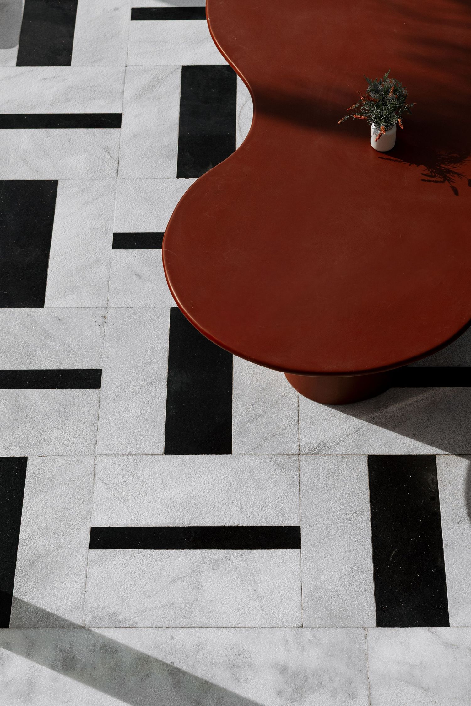 Aerial view of a geometric black and white marble floor with a rust-red organic-shaped table and small potted plant.
